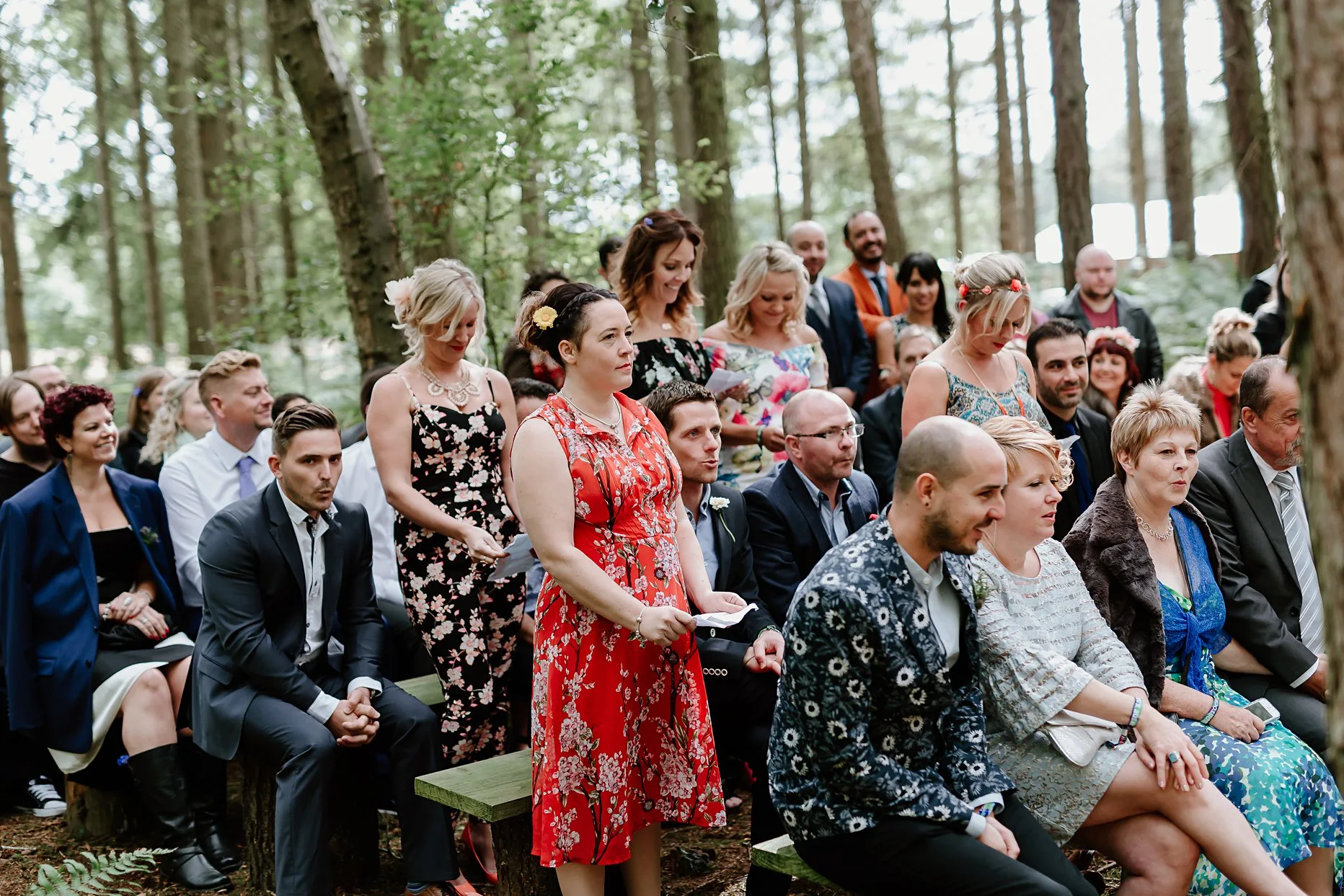 Wedding guests stood up during wedding ceremony singing. The wedding ceremony is outside in the woods at Camp Katur. Guests are surrounded by trees.