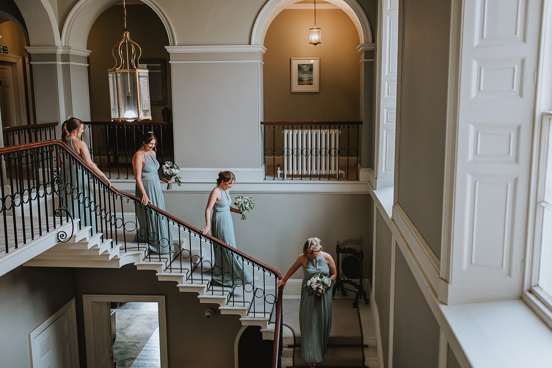 Four bridesmaids dressed in sage green dresses walking down the grand staircase at Saltmarshe Hall Wedding Venue.
