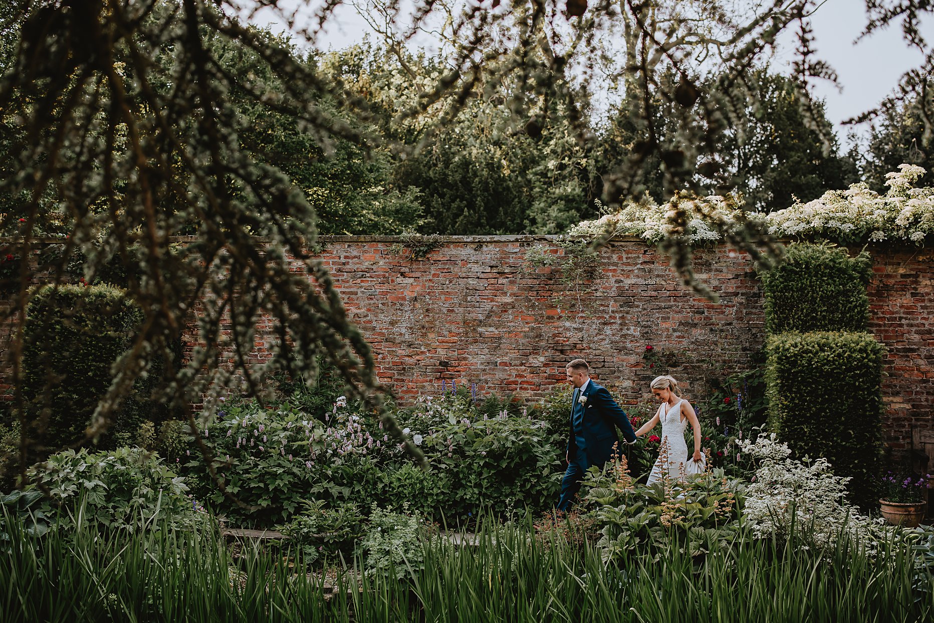 Bride and groom walking hand in hand through the gardens of Saltmarshe Hall. The groom is in front of the bride and leading her. There is a brick wall behind them and they are framed by the foliage and trees in the foreground of the photo.