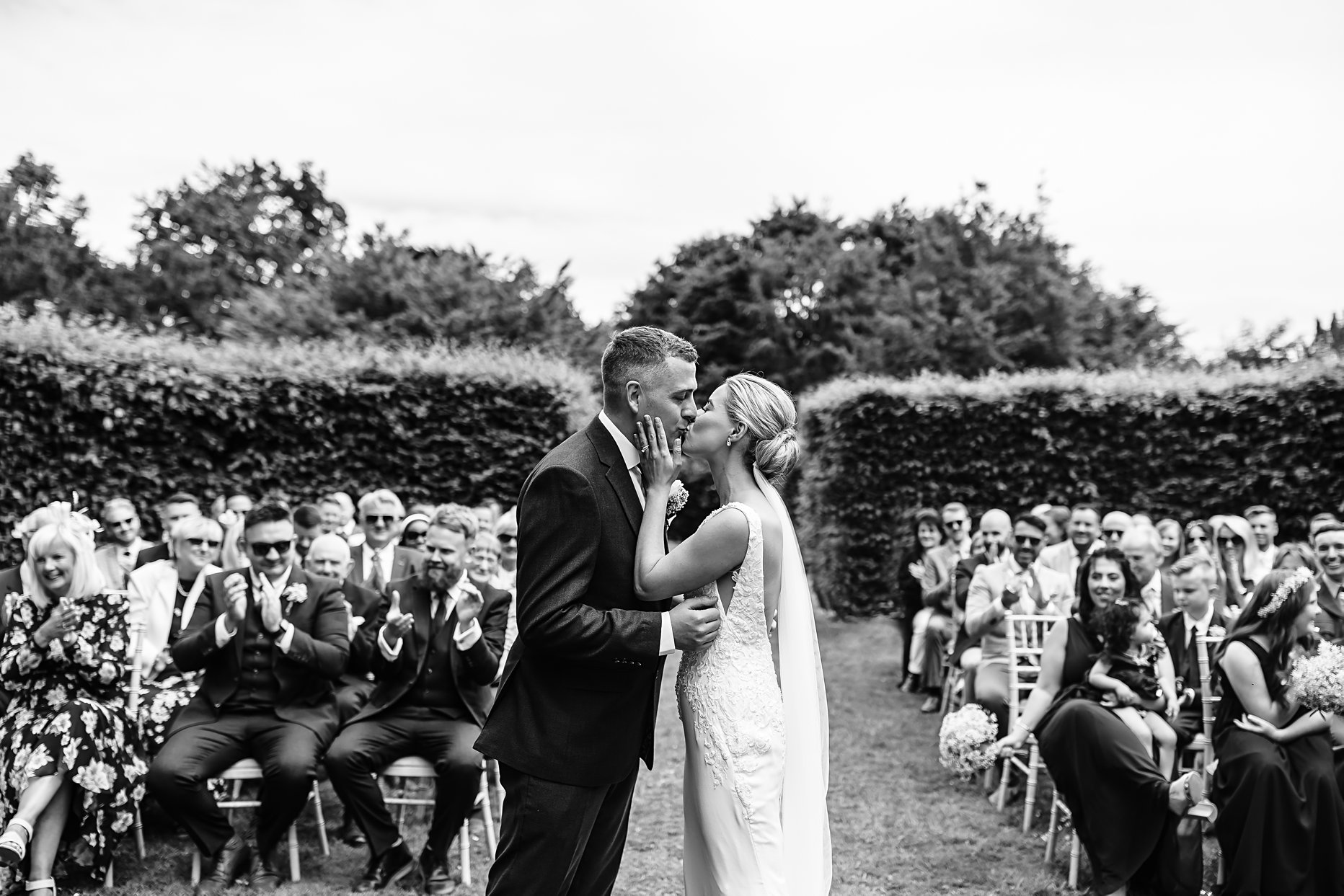 Outdoor ceremony at Saltmarshe hall. Black and white photo of a bride and groom having their first kiss after their ceremony. The bride is holding the grooms face and guests are sat behind clapping. 