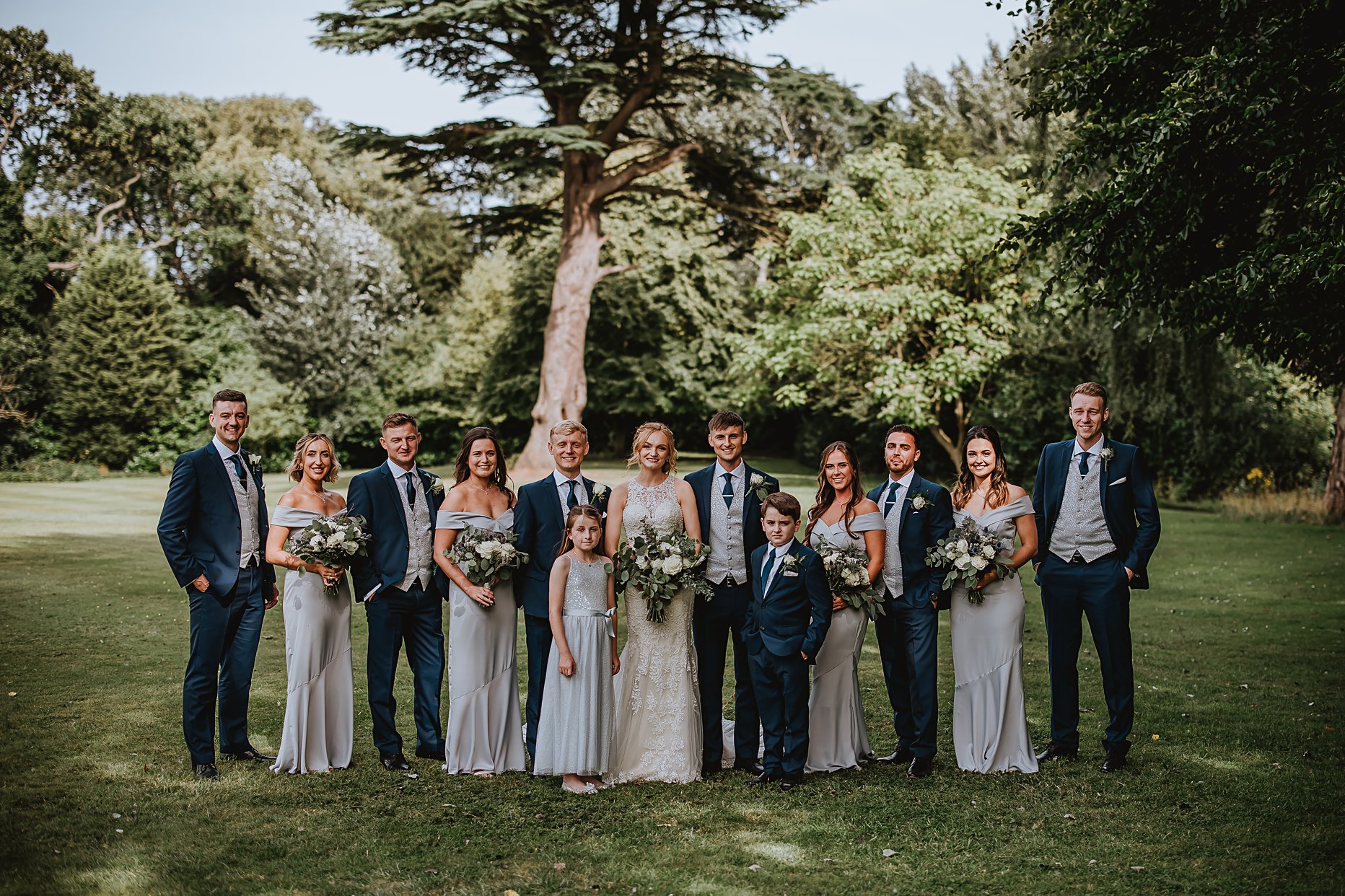 Bride and Groom stood with groomsmen and bridesmaid in the gardens of Saltmarshe Hall. Groom and groomsmen are wearing a navy blue suit and bridesmaids are wearing grey floor length silk gowns. 
