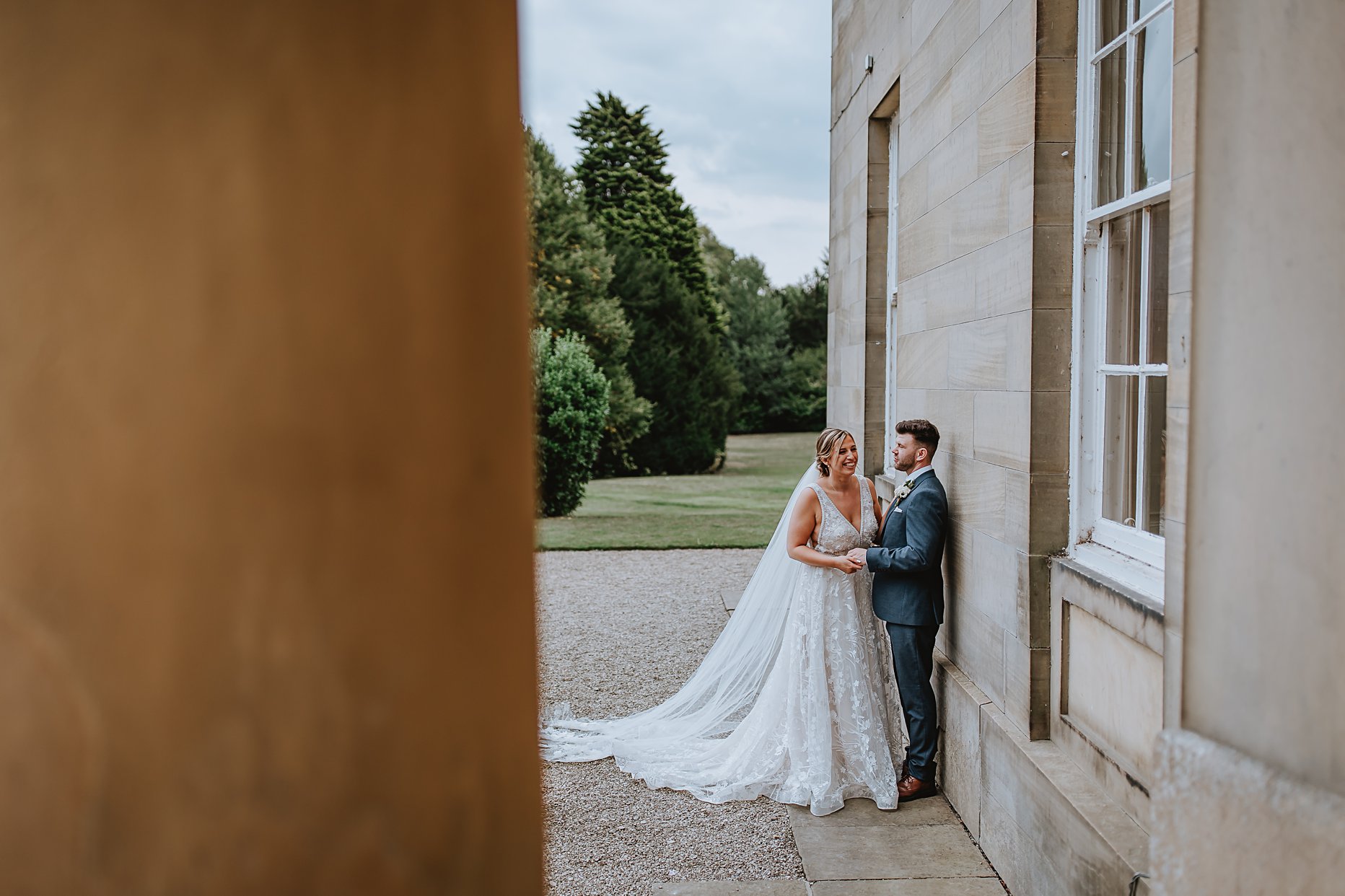 Bride and groom stood against the wall of Saltmarshe Hall. They are holding hands and laughing. Groom is wearing a blue suit and the bride is wearing a fit and flare wedding dress with long veil. 
