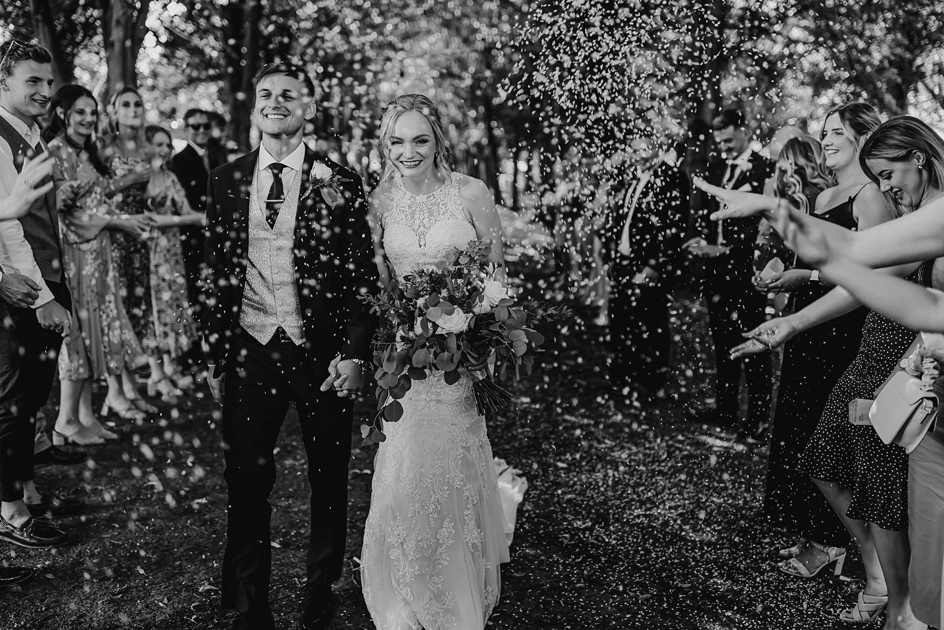 Black and white photograph of bride and groom walking through confetti tunnel at Saltmarshe Hall. They are both smiling whilst guests surround them throwing white confetti discs into the air.