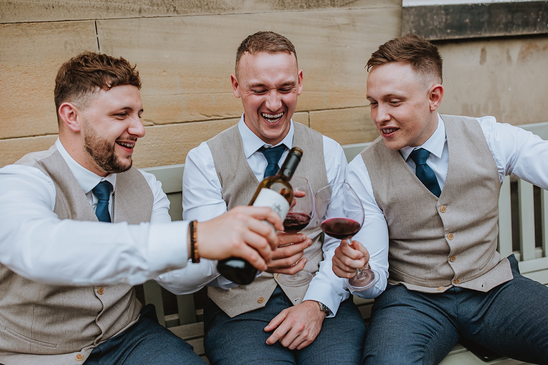 Three groomsmen sat down enjoying a glass of red wine outside Saltmarshe Hall. One groomsman is pouring the red wine while the other two laugh. They are wearing beige waistcoats and blue trousers.