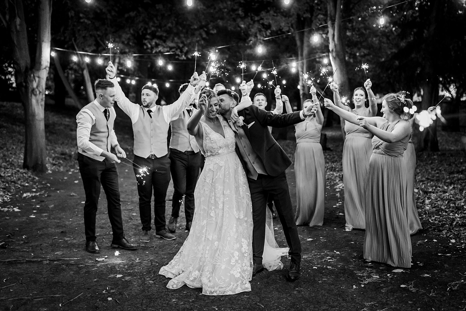 Black and white photograph of a bride and groom holding sparklers. Bridesmaids and groomsmen stand behind them waving the sparklers. Groom is leaned in towards the bride kissing her on the cheek.
