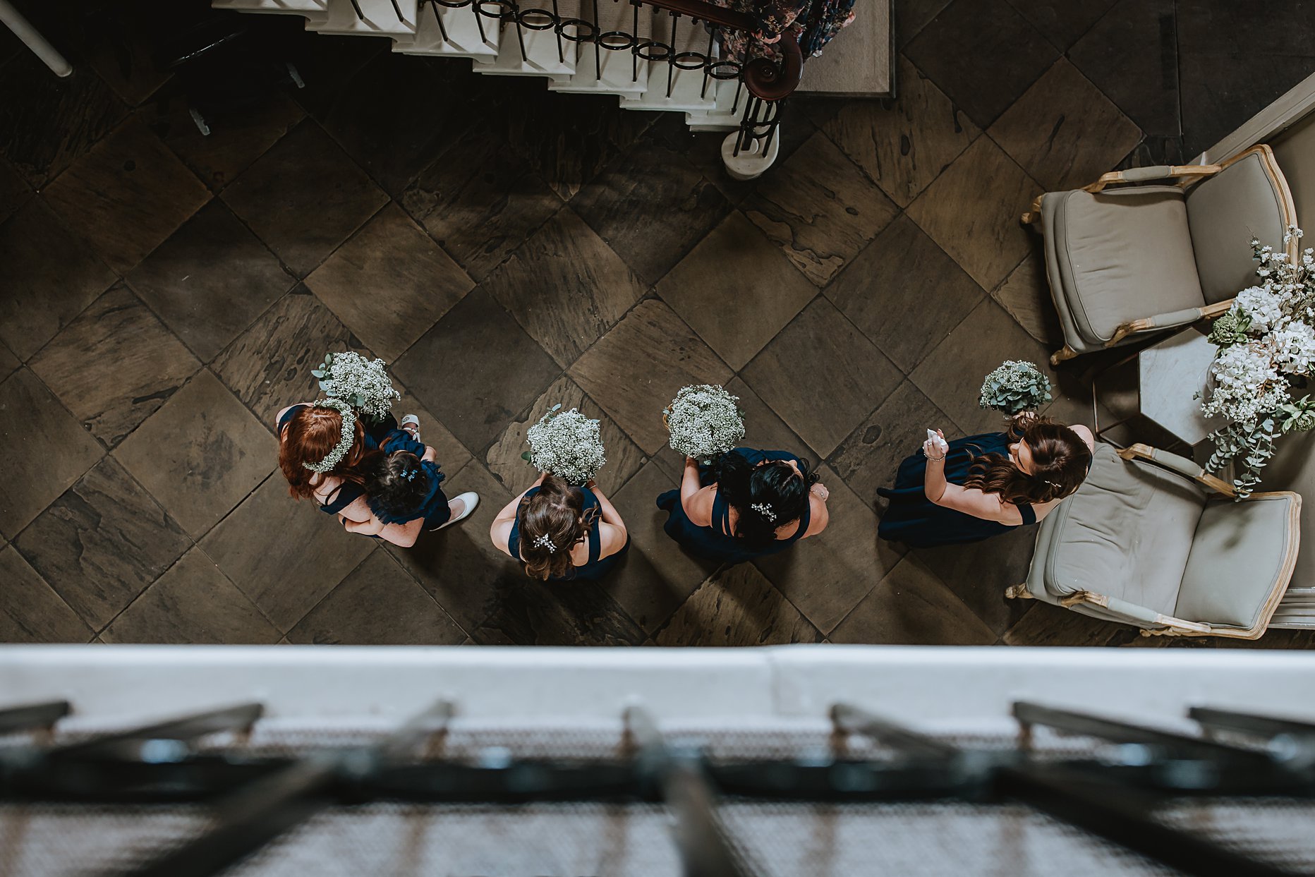 Aerial view of bridesmaids stood in a line at the bottom of the staircase. They are wearing navy blue dresses and holding bouquets of gypsophila. 