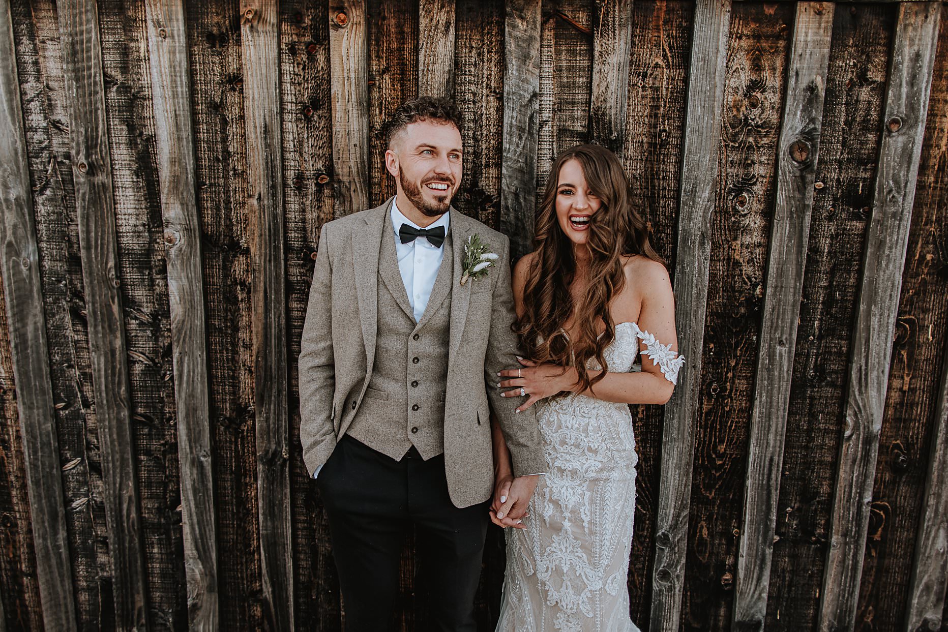 Bride and Groom leaning against wooden panelled wall. They are holding hands and laughing towards the camera. Bride has long brown hair and groom is wearing a bow tie.