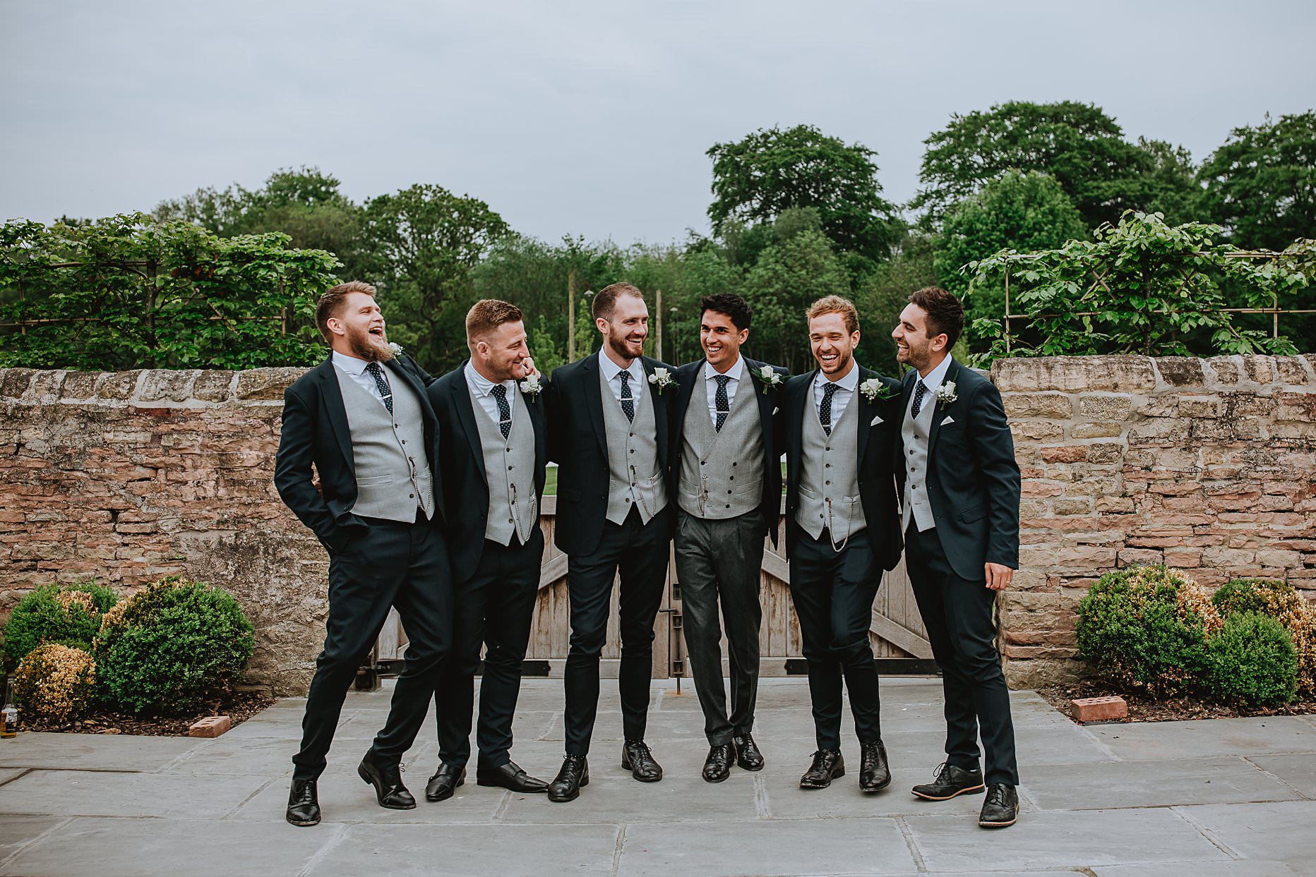 Groom and his groomsmen stood near a brick wall laughing and chatting. Groomsmen wear navy suits and a grey waistcoat.