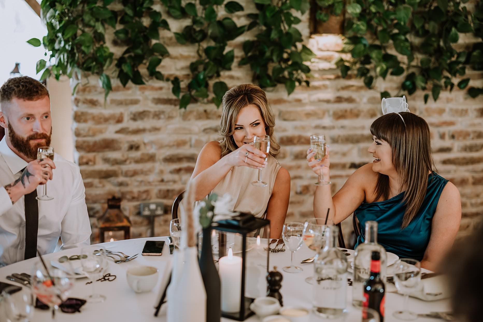 Wedding guest sat at table during wedding breakfast. She is raising a glass of prosecco to toast the bride and groom.