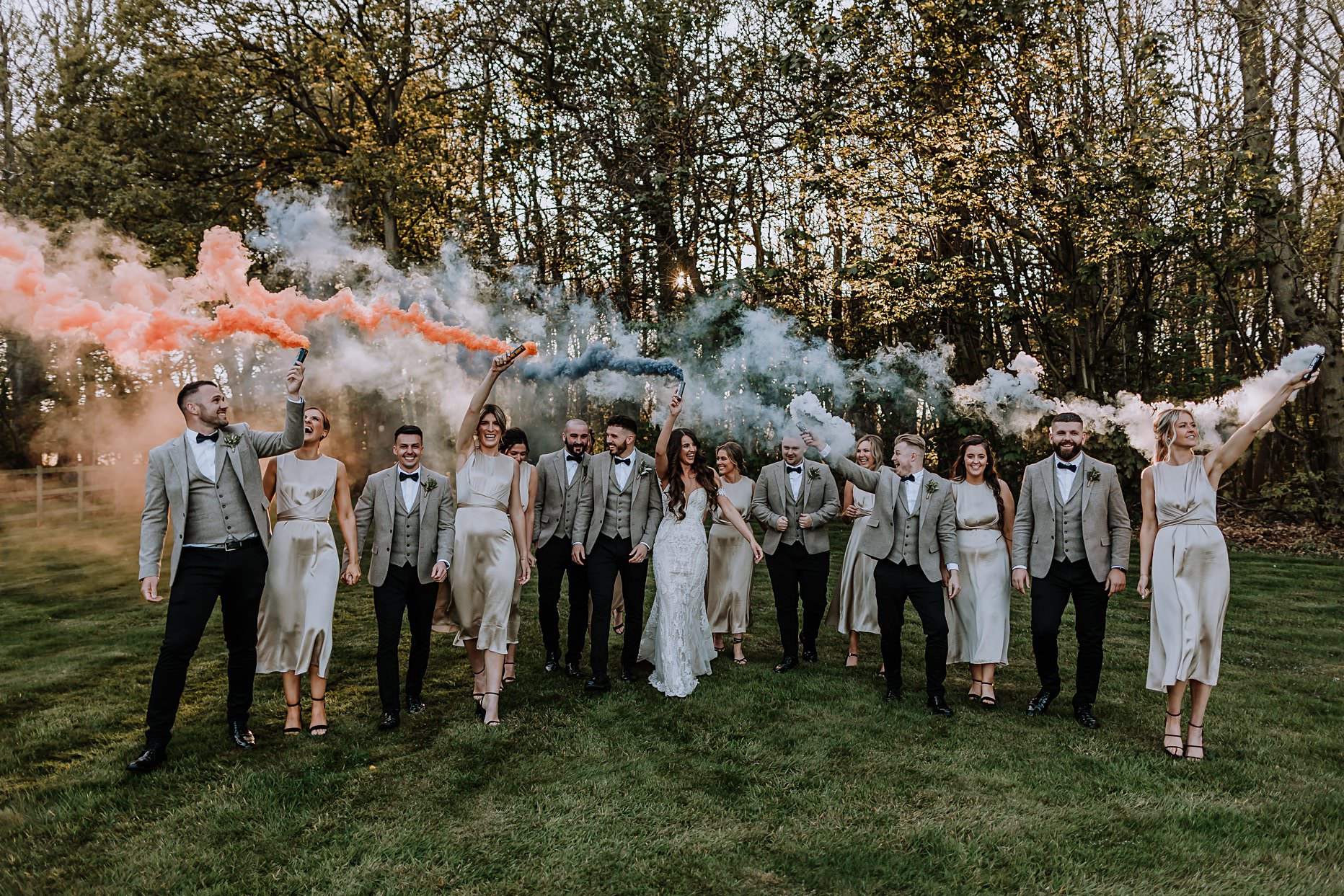 Bride and groom walking with bridesmaids and groomsmen holding smoke bombs in the air. They are walking on the garden at Hazel Gap Barn.