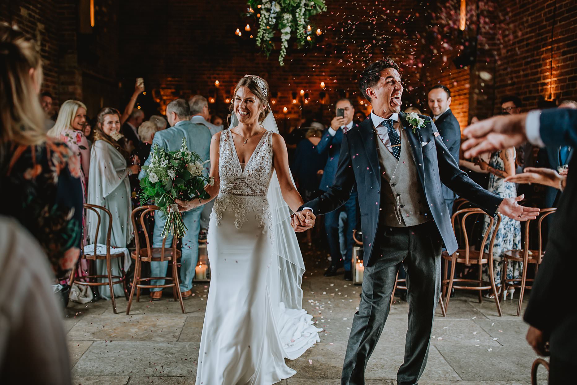 Bride and groom walking down the aisle whilst guests throw confetti at them. They are both smiling and groom is making a silly face.