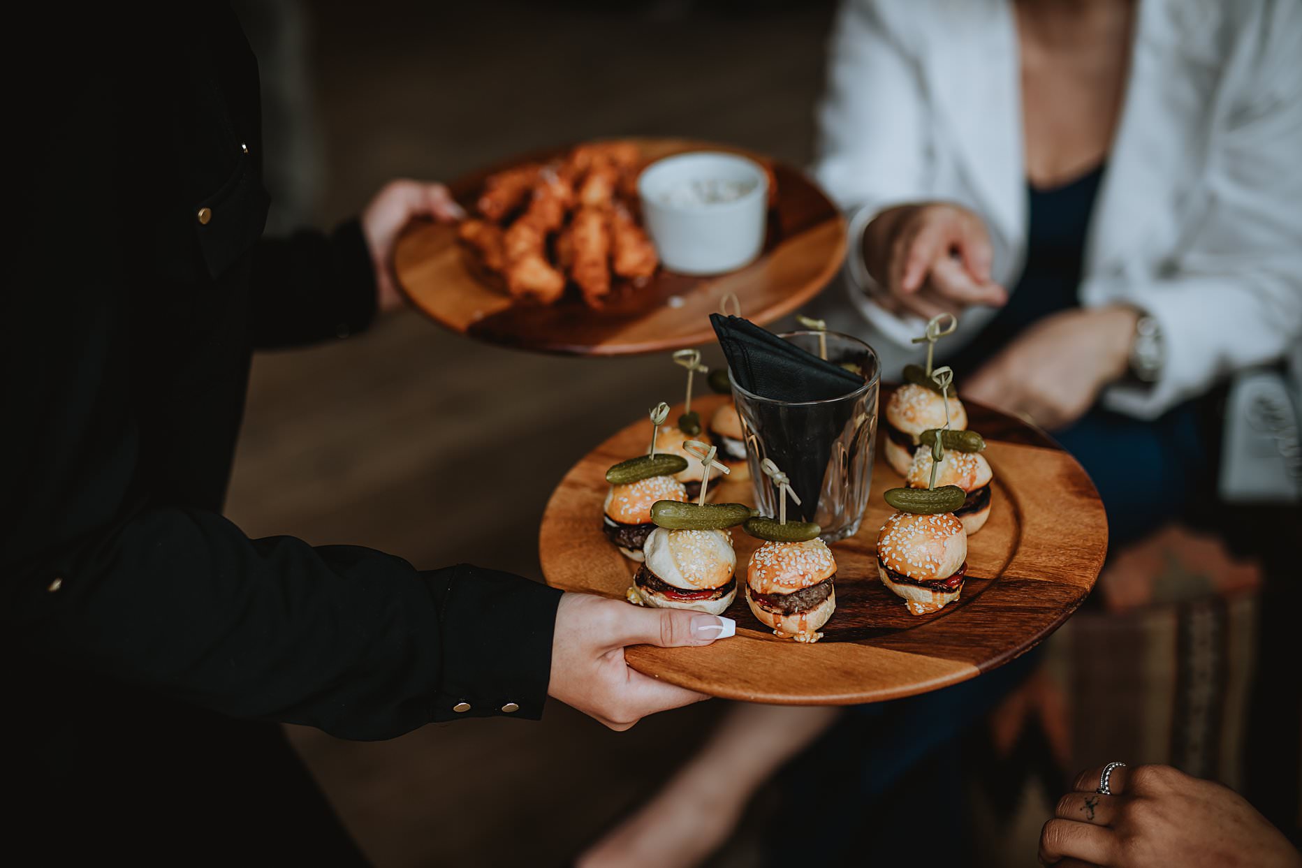 Waitress handing out wedding canapés to guests. The canapés are mini hamburgers with pickles and are presented on a wooden board.