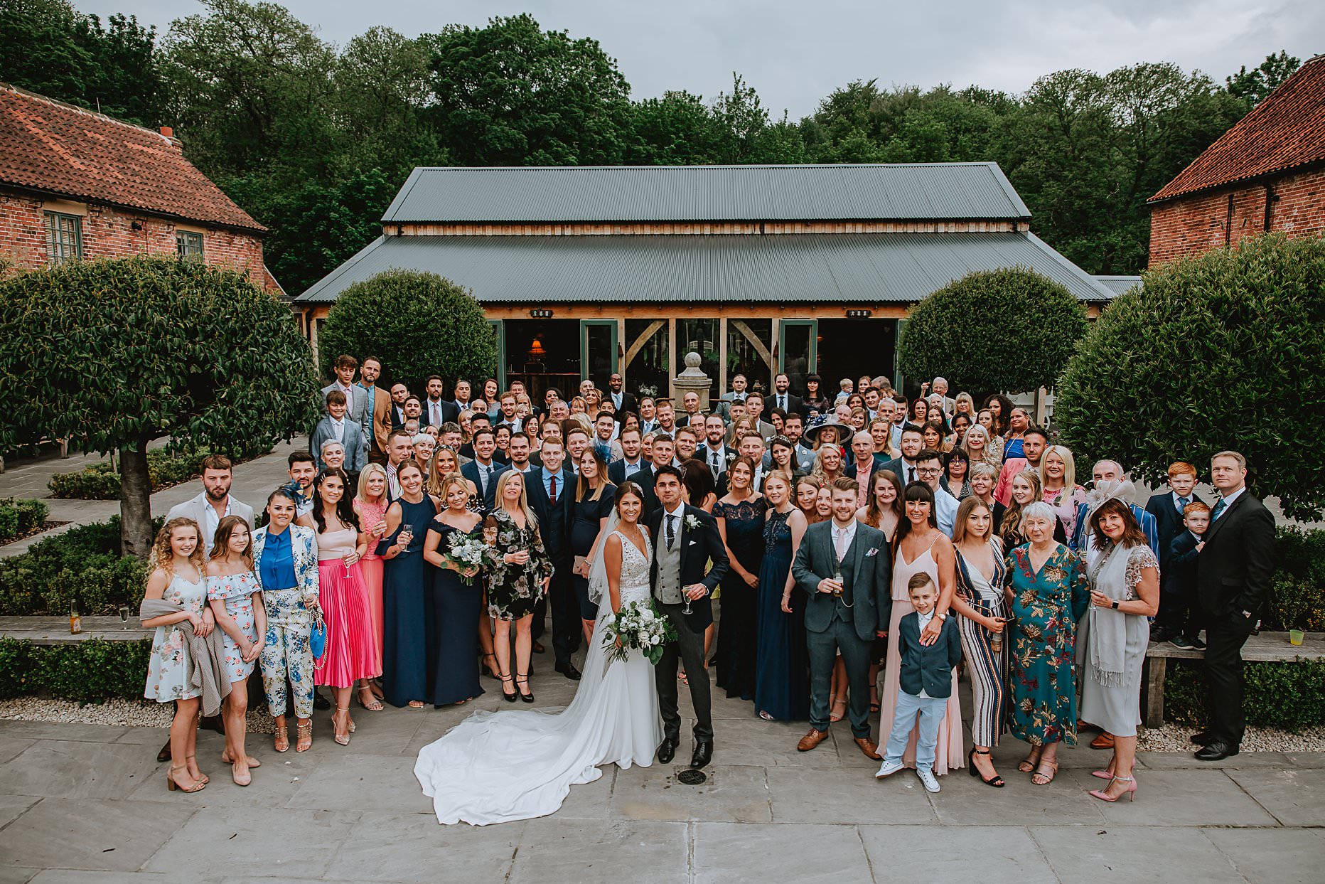 A large group photo of all wedding guests outside Hazel Gap Barn wedding venue. Bride and groom are stood at the front of the group.
