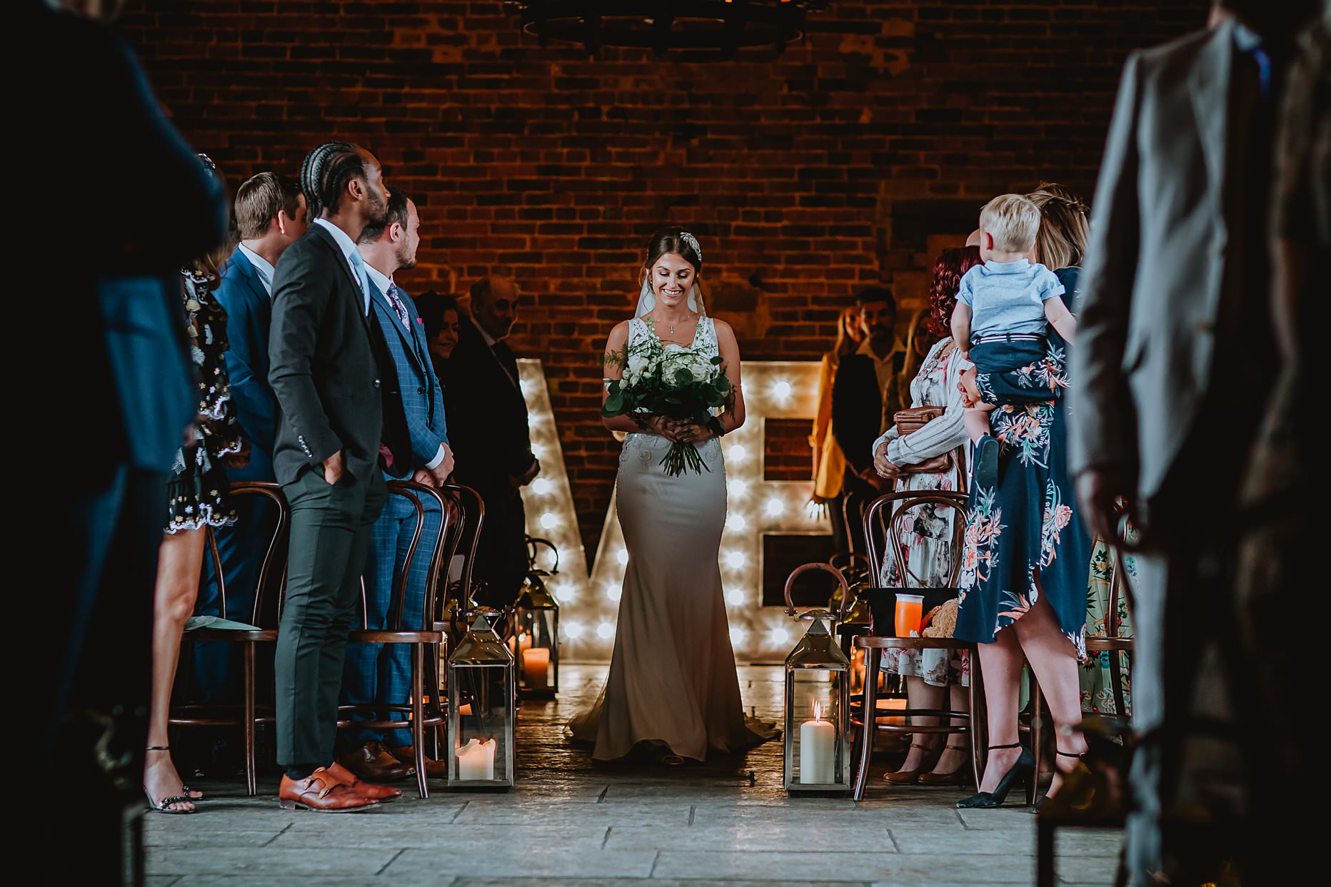 Bride walking down the aisle on her own at Hazel Gap Barn. She is holding a bouquet and guests are stood up either side of her watching her walk.
