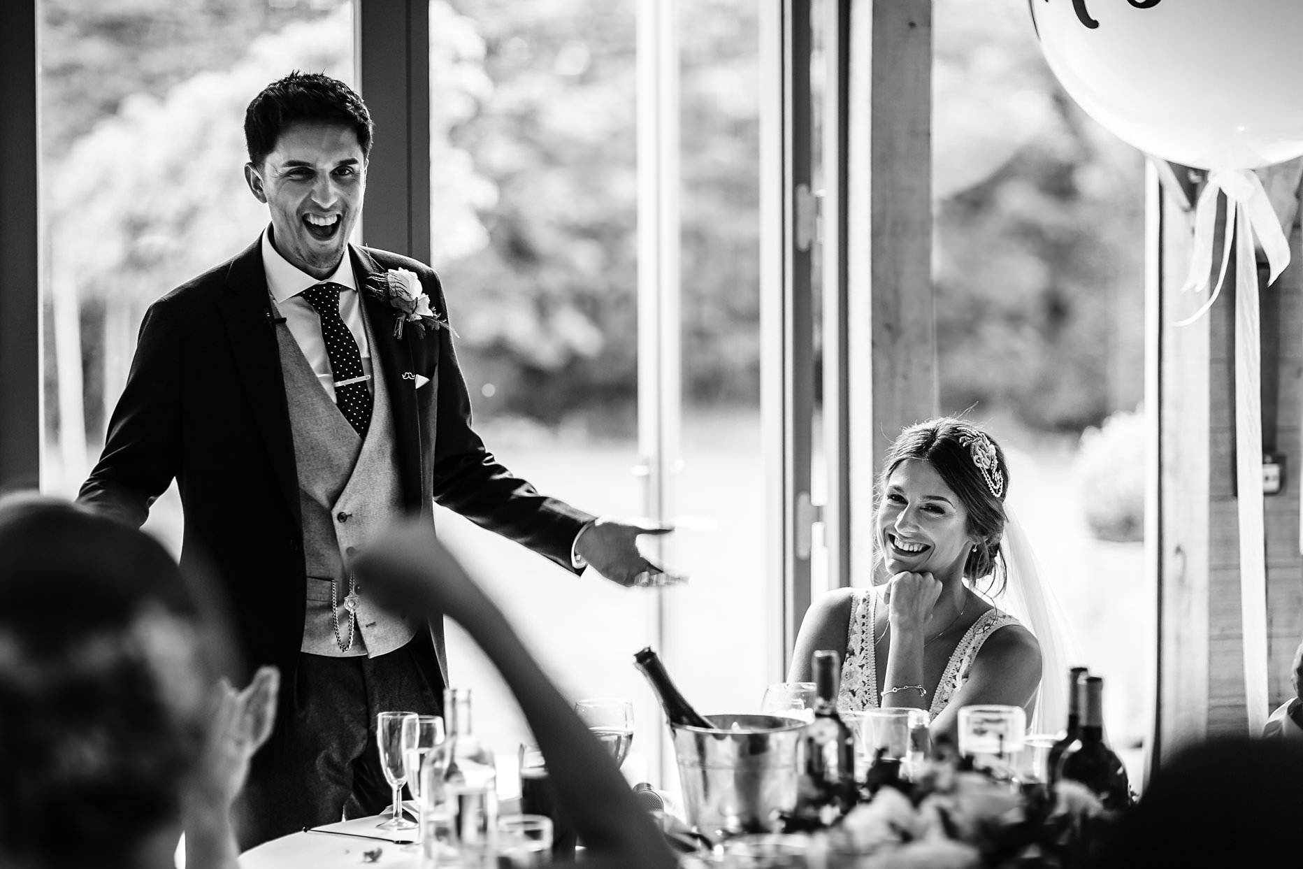 Groom delivering a speech on his wedding day. His wife is sat next to him laughing. Photograph taken at Hazel Gap Barn.