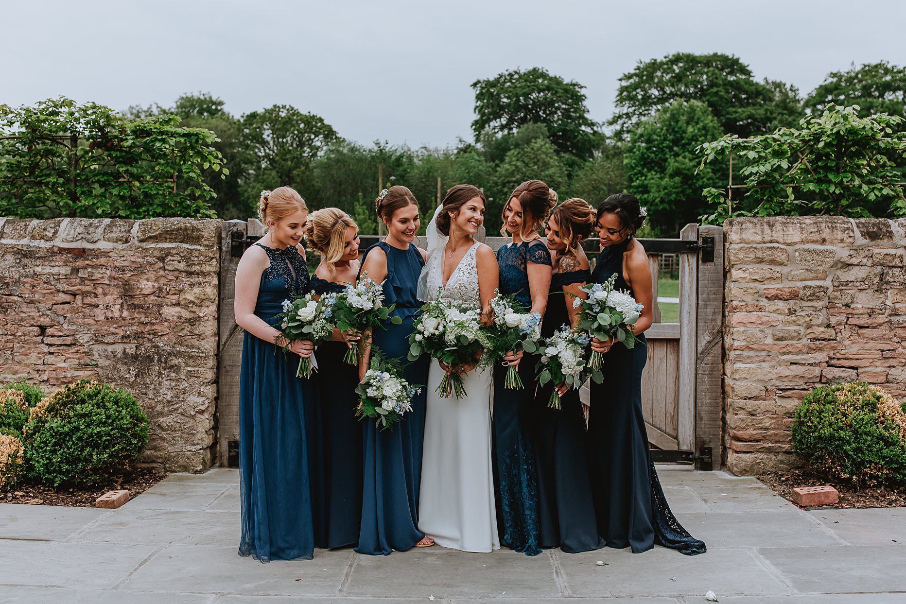 Bride and her bridesmaids stood near a brick wall talking and laughing. They are all holding bouquets of green foliage and white flowers.