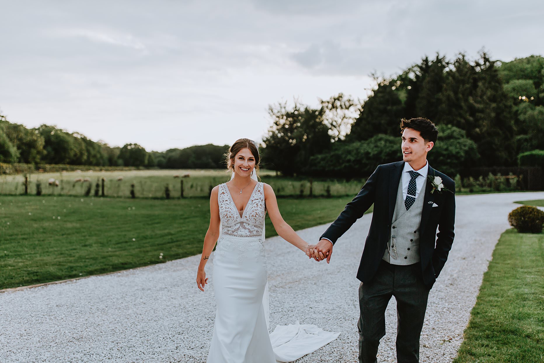 Bride and groom walking hand in hand at the front of Hazel Gap Barn. The bride is wearing a floor length wedding dress and is happy and smiling towards the camera. The groom is wearing a navy suit and looking into the distance.