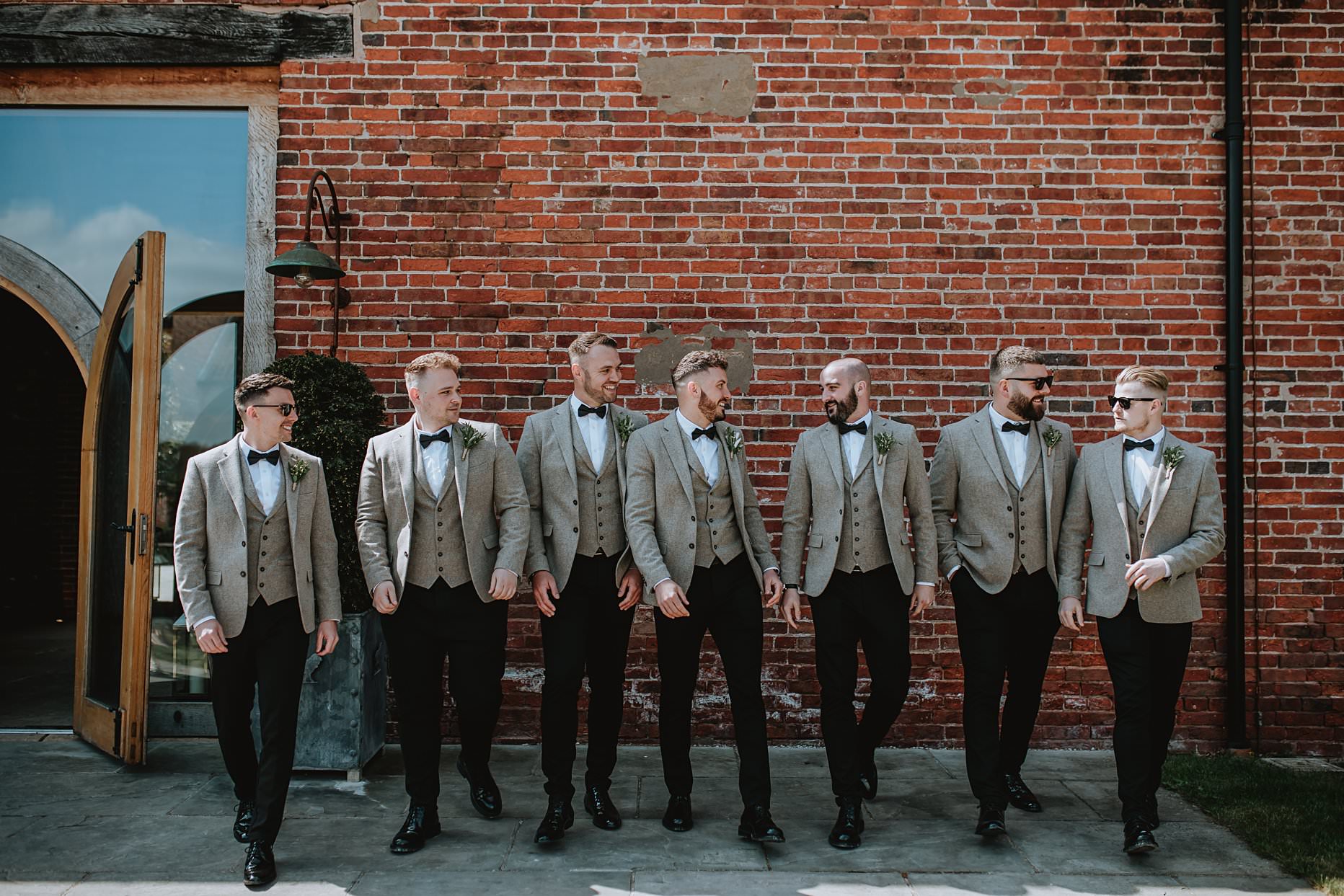 Groomsmen walking towards the camera laughing and chatting to each other. Groomsmen are wearing beige suit jackets, black trousers and bow ties.