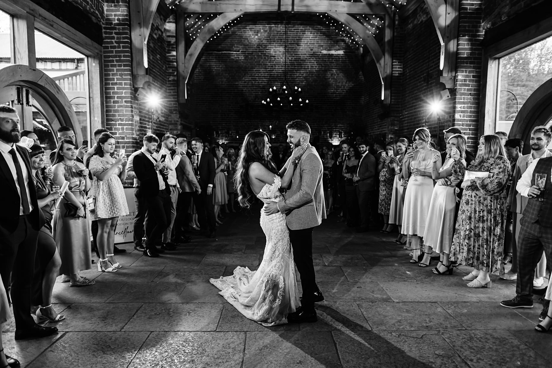 Bride and groom having their first dance at Hazel Gap Barn. Wedding guests surround them. They are dancing in the centre of the room and two bright lights shine in the background.