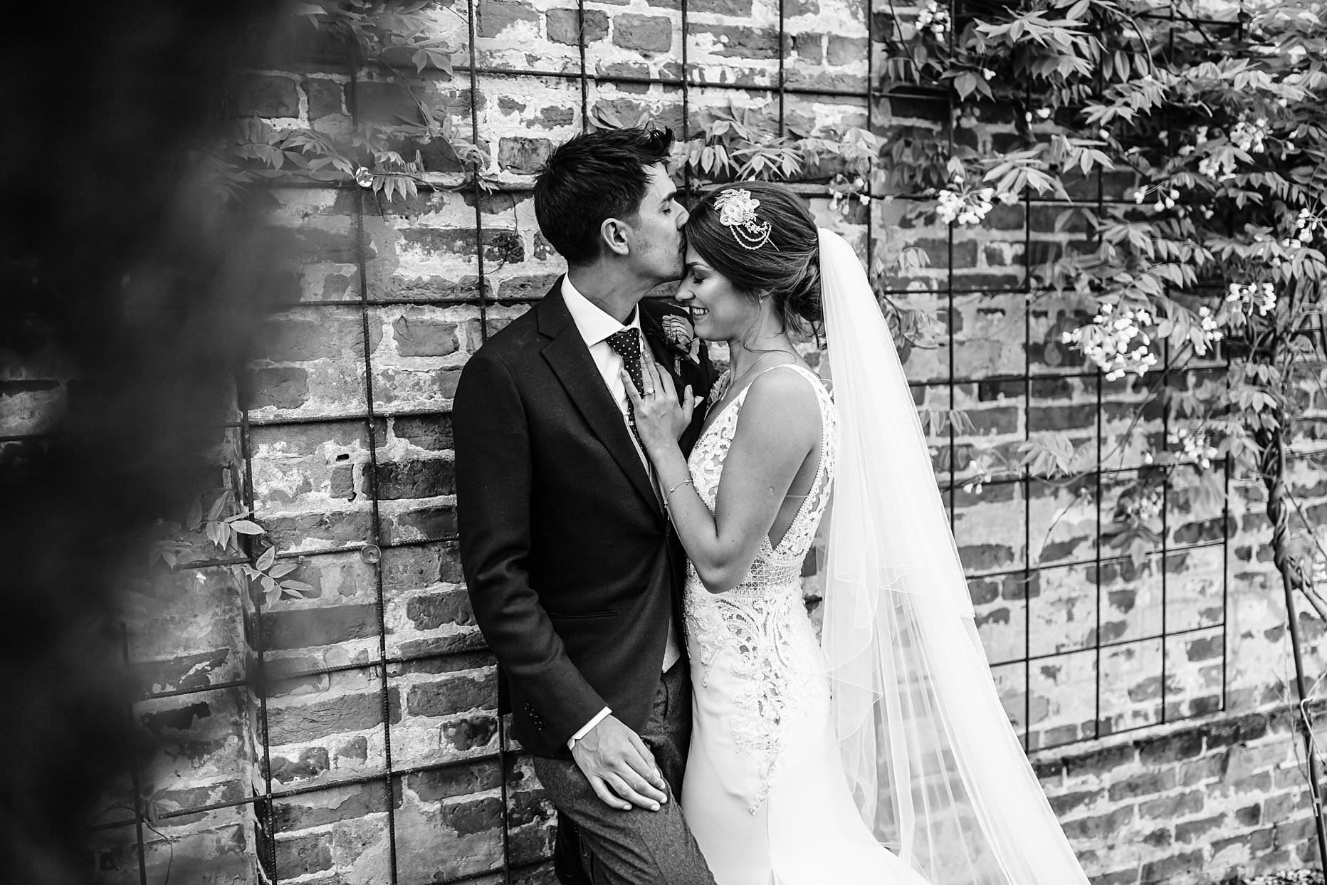 Groom is rested against a brick wall, his wife is leaned into him and he is kissing her forehead.