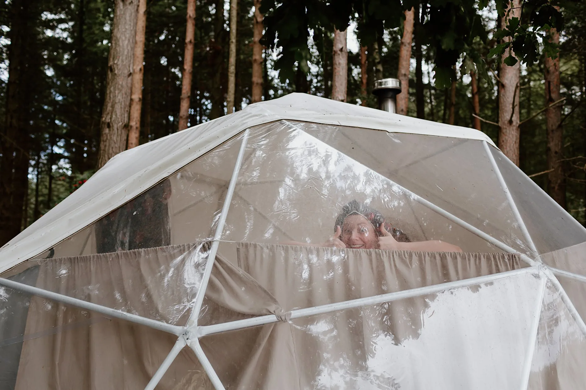 Wedding guests inside glamping shower pod with shower cap on and giving two thumbs up.