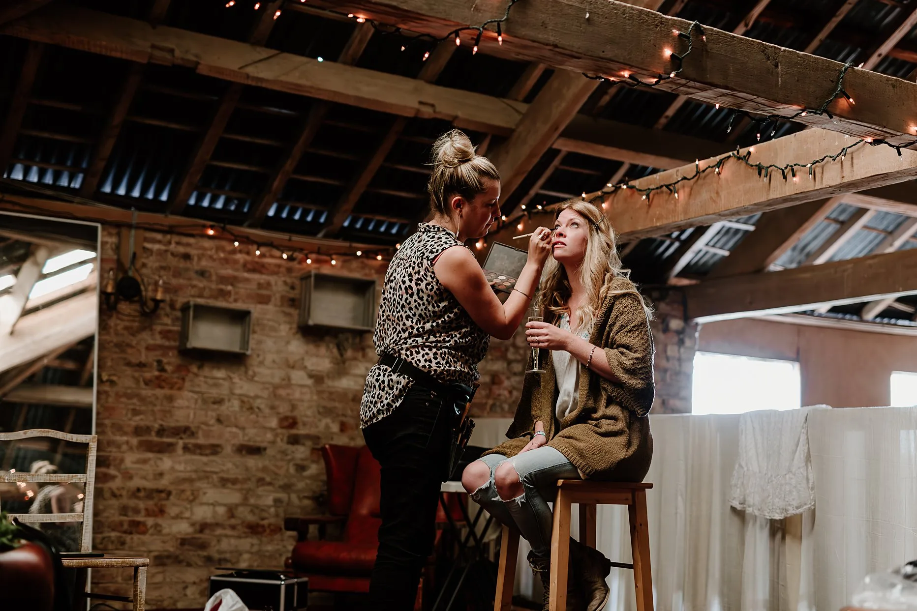 Bride having her make-up done before her wedding at Camp Katur. She is sat on a stool with a glass of champagne. 