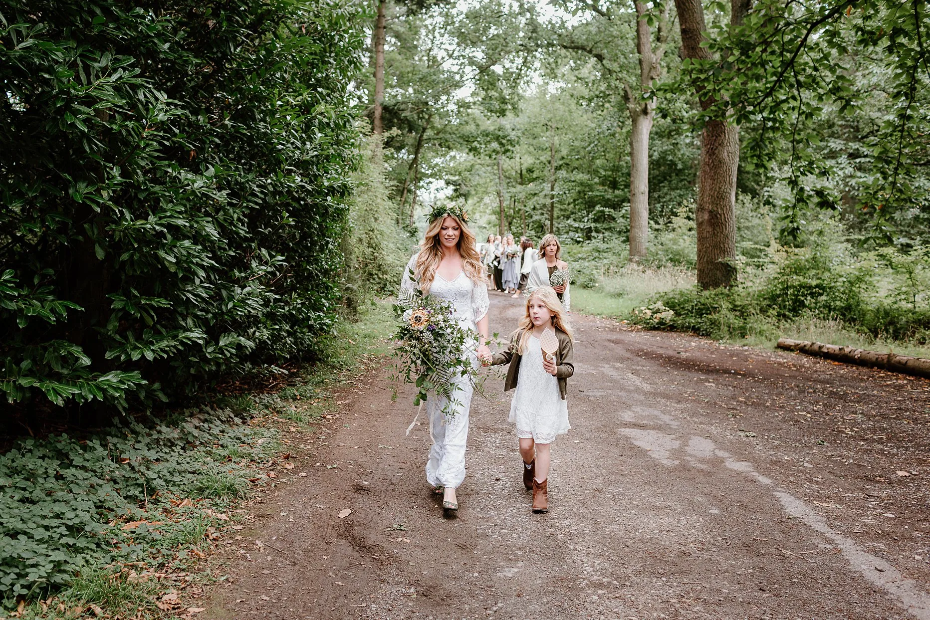 Bride holding hands with small flower girl walking through the woods to outdoor ceremony at camp katur. Bride is wearing a long white crochet wedding dress and holding a large wild foliage bouquet.