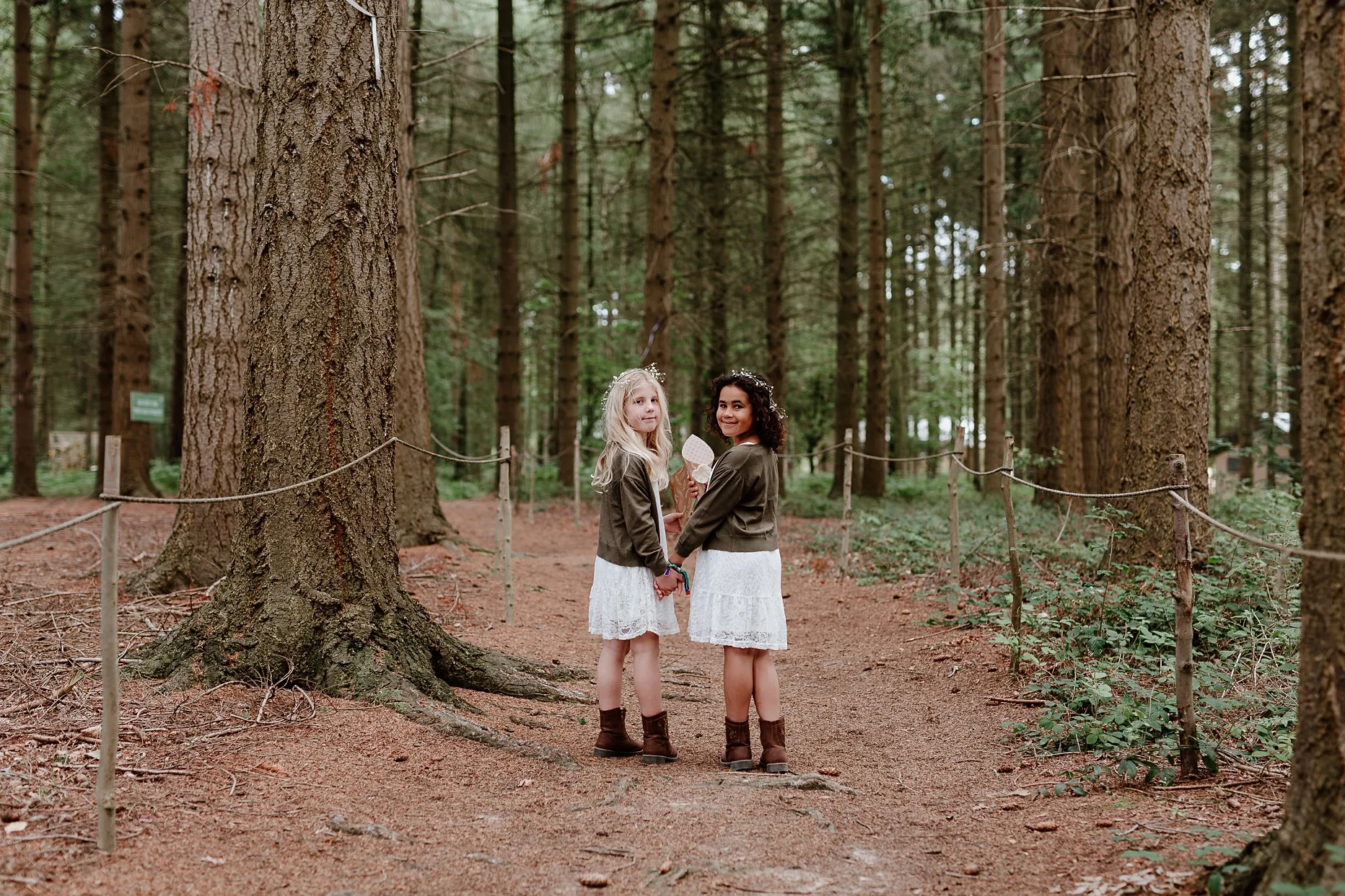 Two young flowergirls walking through the woods to outdoor wedding at camp katur. Flowergirls are wearing short white lace dresses with brown cowboy boots.