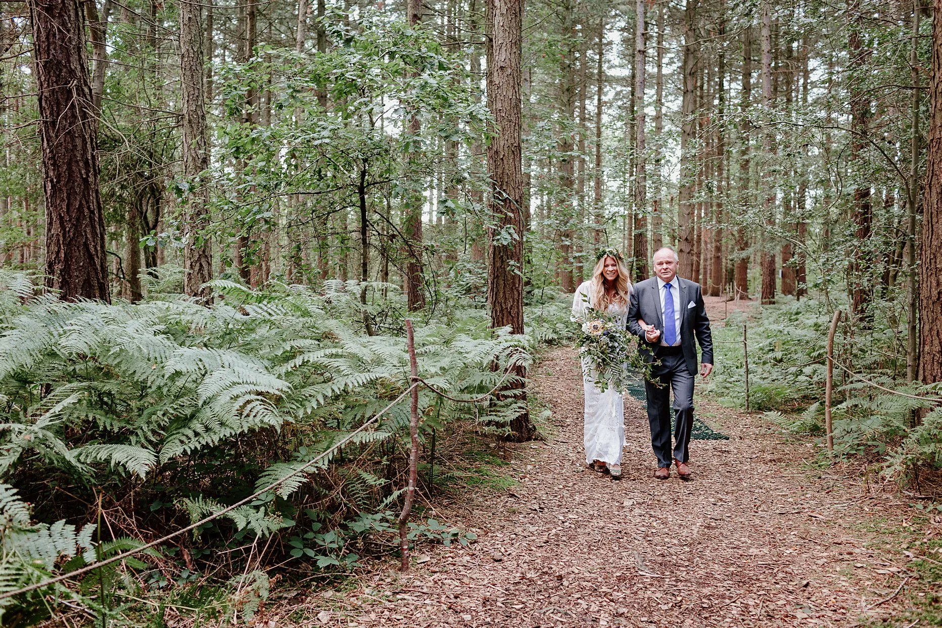 Bride walking with her dad through the woods towards the woodland chapel ceremony area at Camp Katur