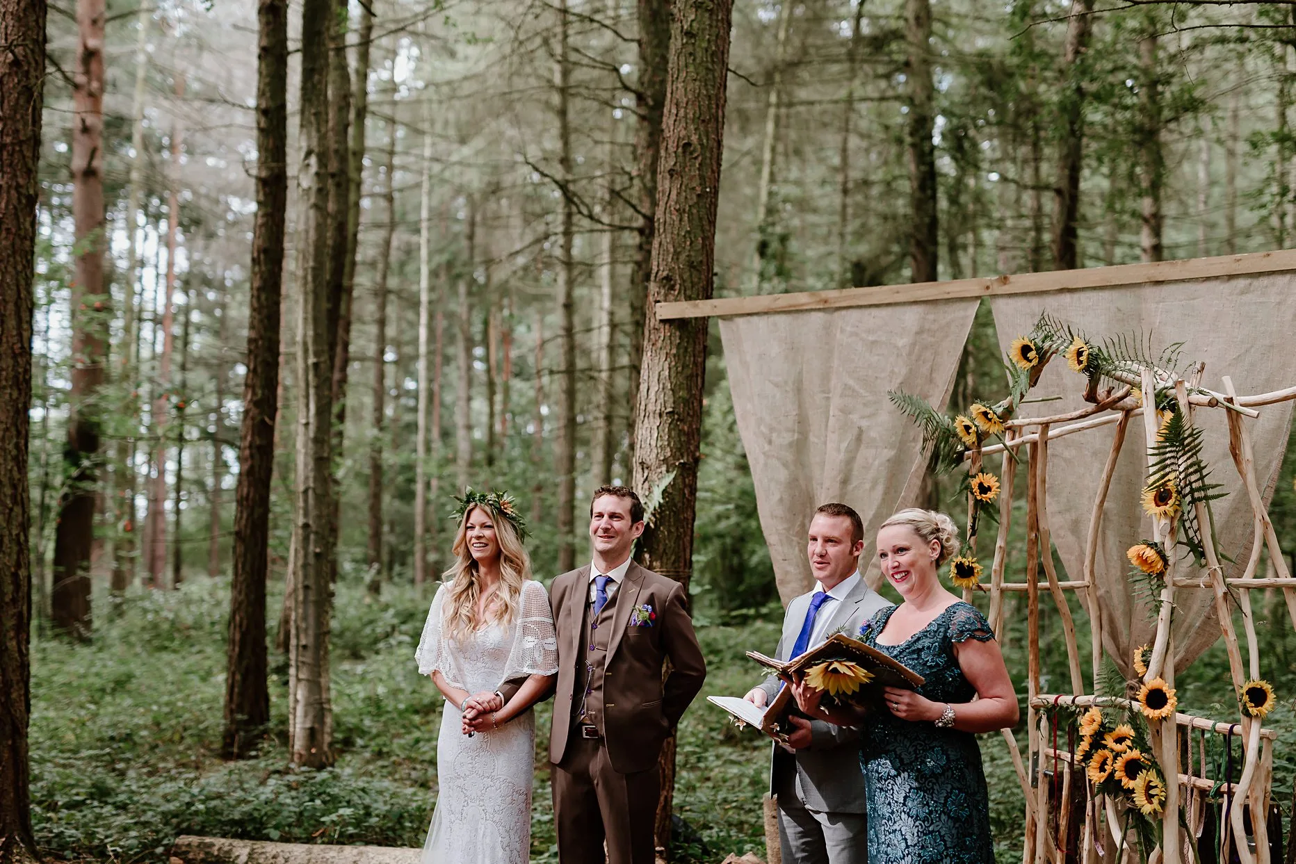 Bride and groom stand with two celebrants during their wedding ceremony in the woods at Camp Katur. They are stood next to a archway of sunflowers.