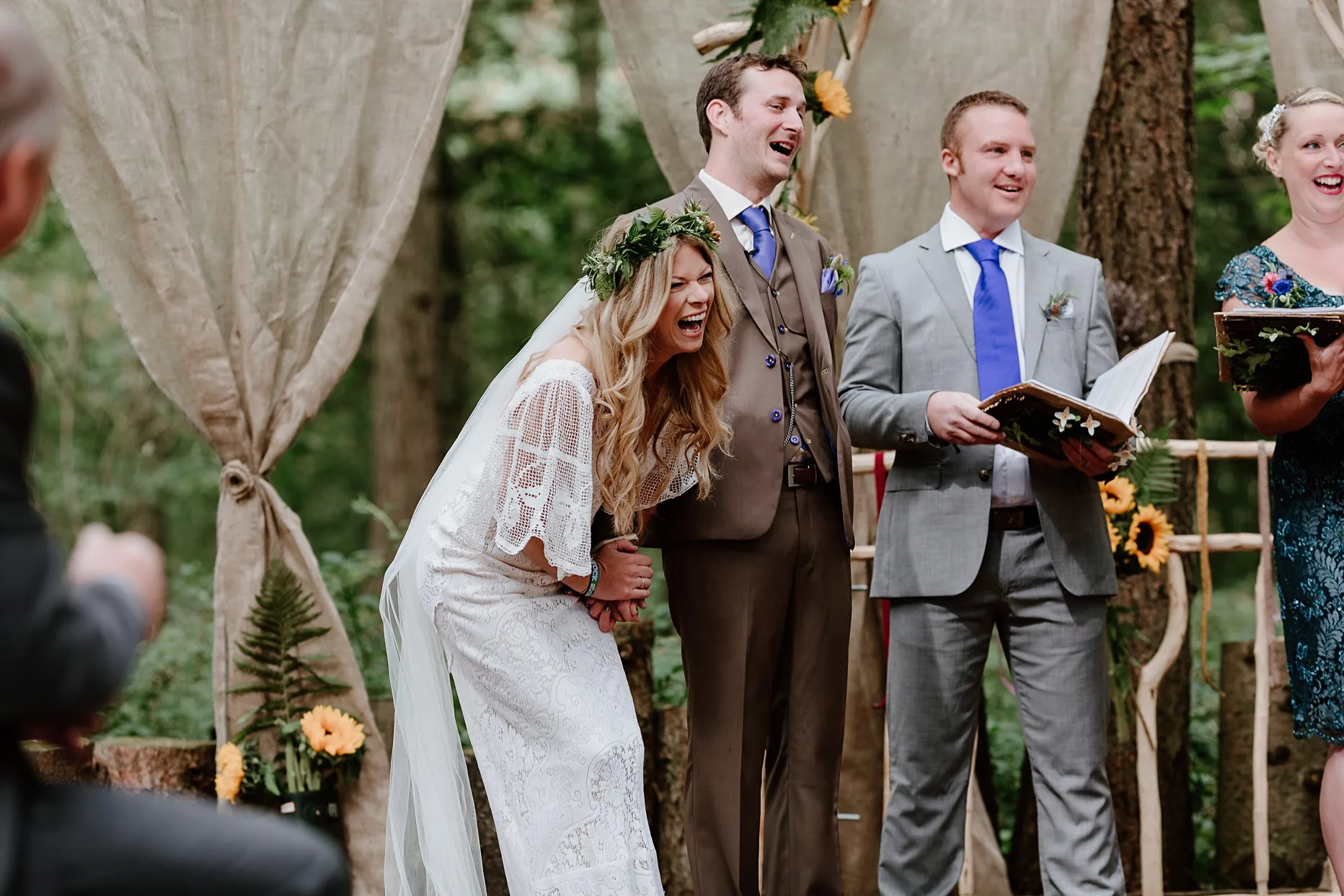 Bride and groom holding hands laughing during their wedding ceremony in the woods. Bride is wearing a boho crochet white wedding dress and flowercrown. The ceremony is outside in the woods at the woodland chapel, Camp Katur.