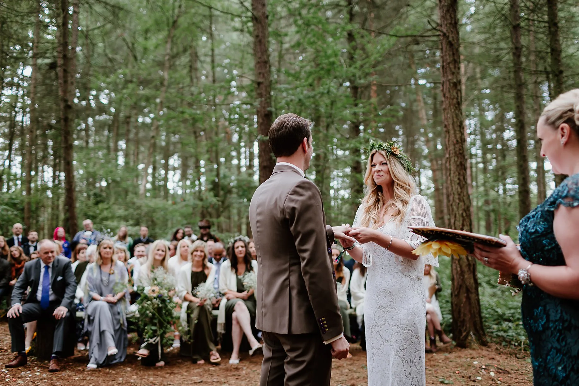 Bride and groom exchanging rings during their wedding ceremony in the woods. They are surrounded by trees and wedding guests are seated in the background watching them and smiling.
