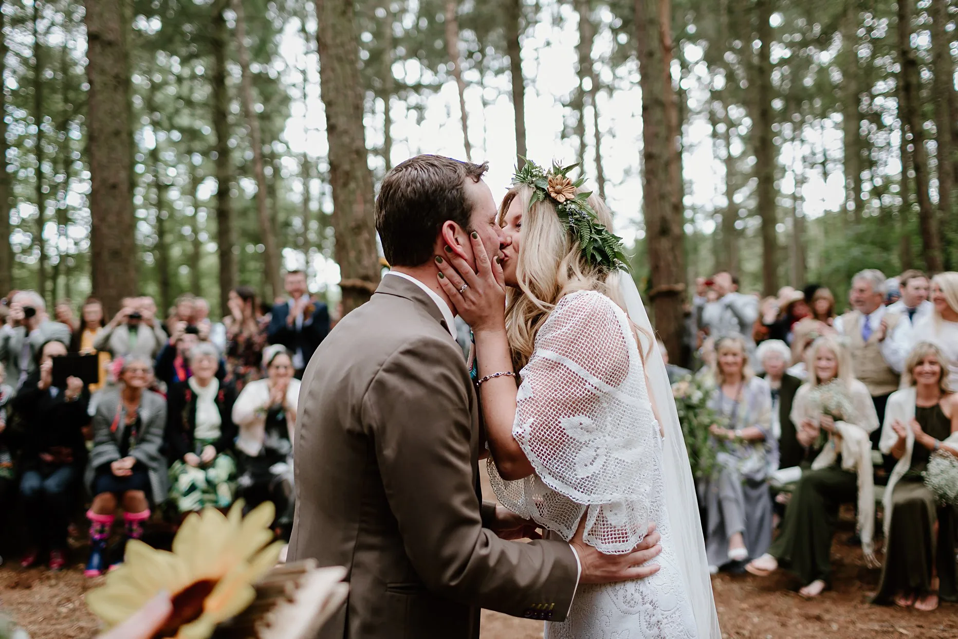 Bride and groom having their first kiss    during outside ceremony in the woods. Wedding guests are in the background clapping and everyone is surrounded by trees.