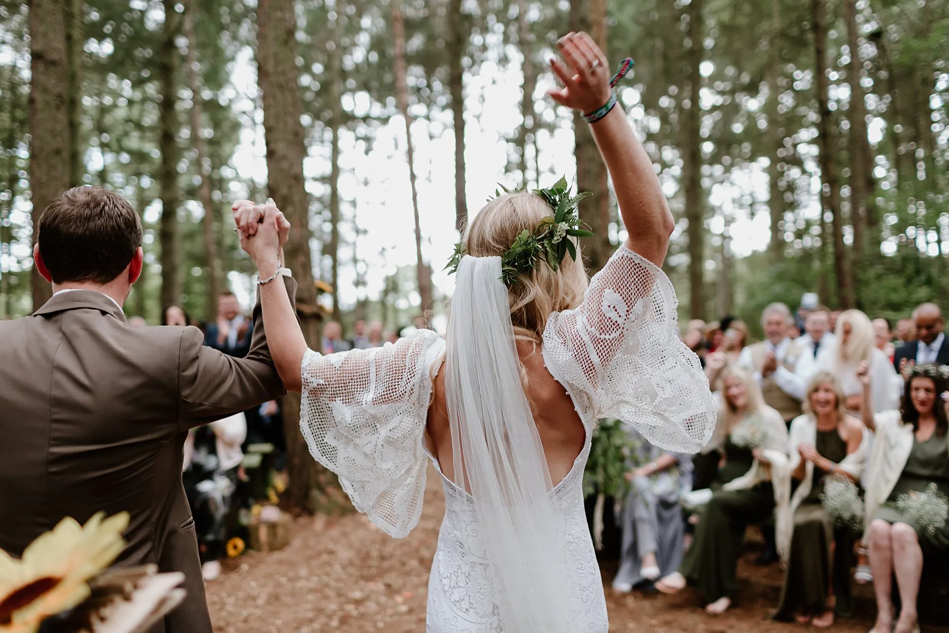 Bride and groom celebrating getting married in woodland wedding ceremony. Bride is throwing up her arms in excitement and looking towards her friends and family who are clapping.