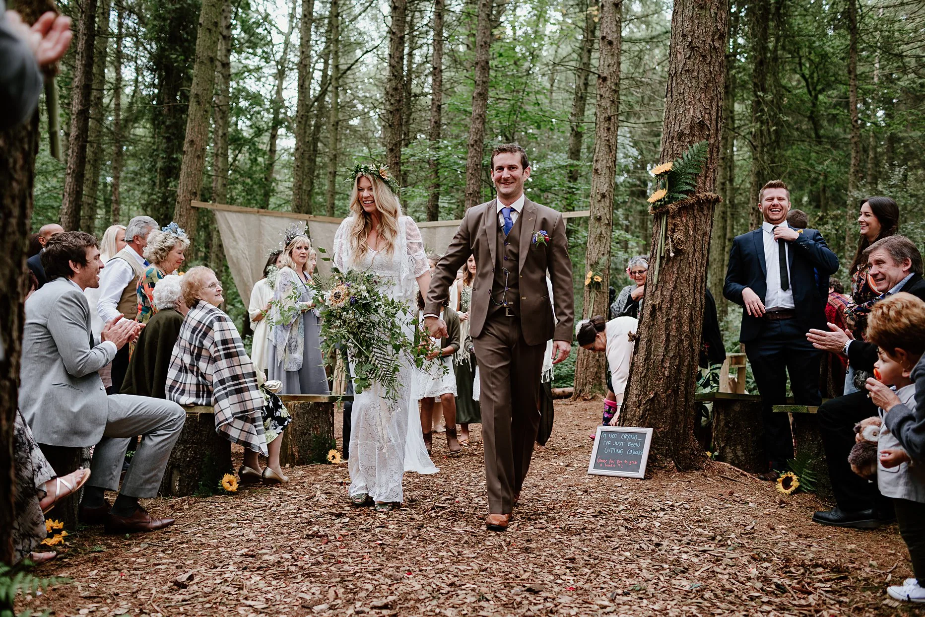 Bride and groom walking back down the aisle after outdoor wedding ceremony at the woodland chapel, Camp Katur. Guests are clapping and smiling and everyone is surrounded by trees.