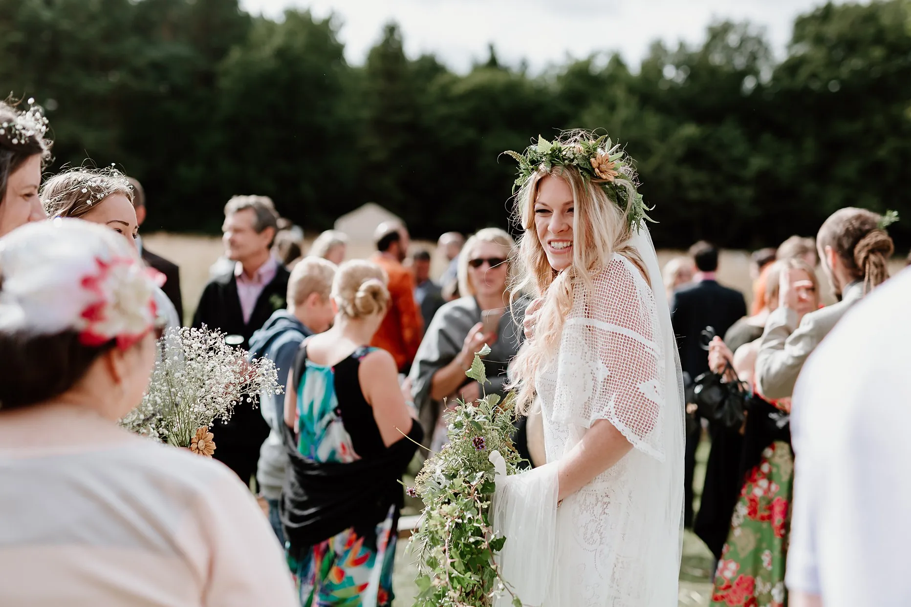 Natural photograph of bride smiling at wedding guests during the drinks reception. Bride is wearing a relaxed white crochet wedding dress and green foliage flower crown.