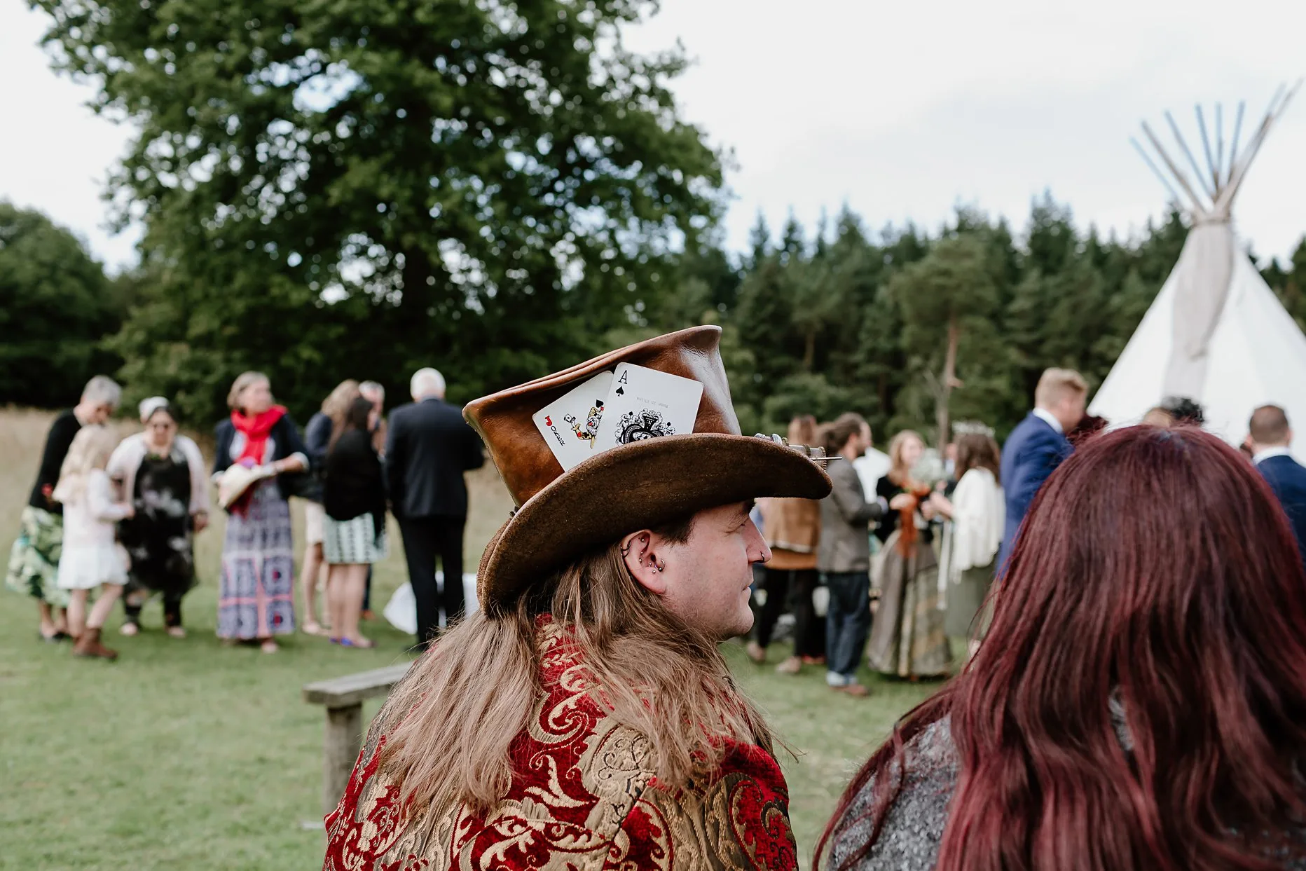 Alternative wedding guest with long hair and wearing a large hat. He is sat down in the meadow at Camp Katur during the wedding drinks reception.