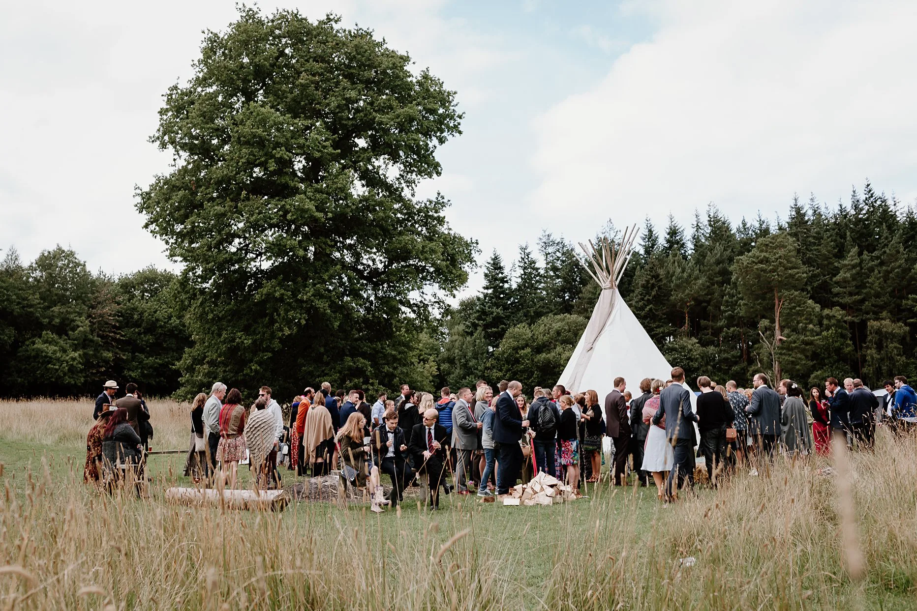 Large group of wedding guests enjoying drinks reception on the meadow at Camp Katur. The meadow is surrounded by tall grass and trees.