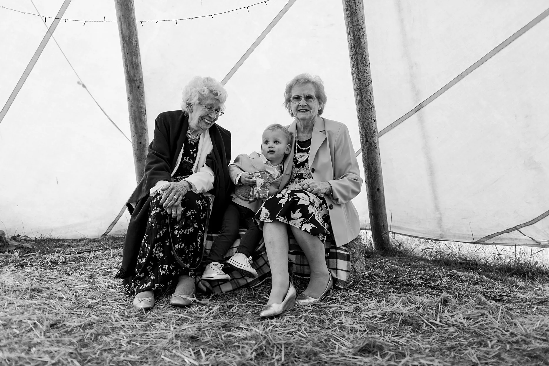 Two older ladies sat on a hay-bail inside a teepee cuddling their grandchild.
