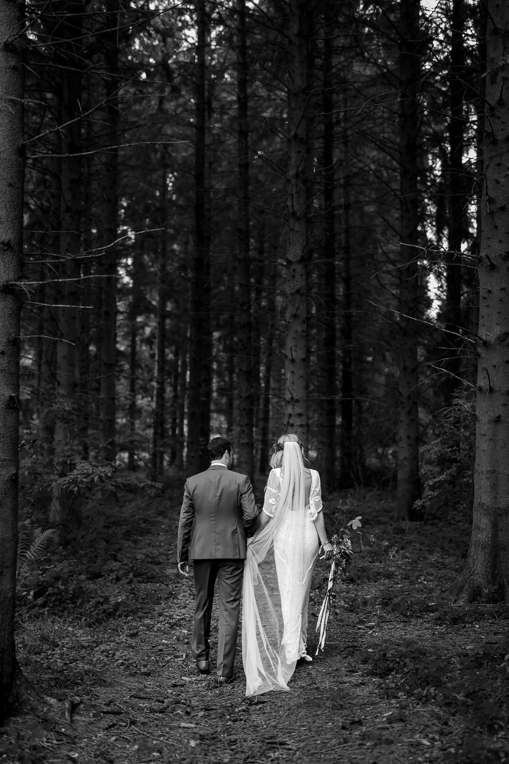 Black and white photograph of a bride and groom walking away from the camera and into the woods. They are holding hands. The bride is wearing a long white veil and is holding a large wedding bouquet. They have just been married at Camp Katur