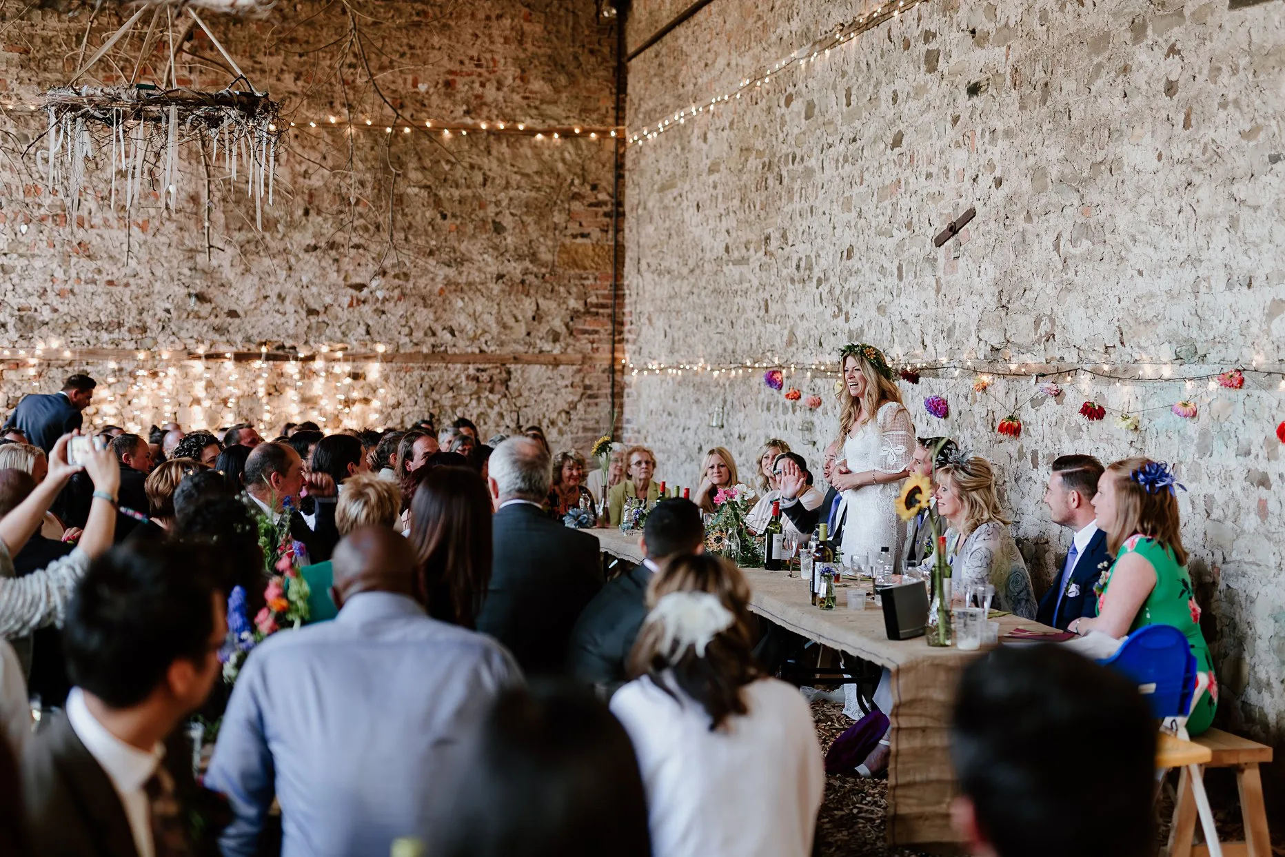 Bride giving a speech at her wedding. She is stood at the top table addressing her friends and family. The speeches are taking place inside an old barn at Camp Katur.