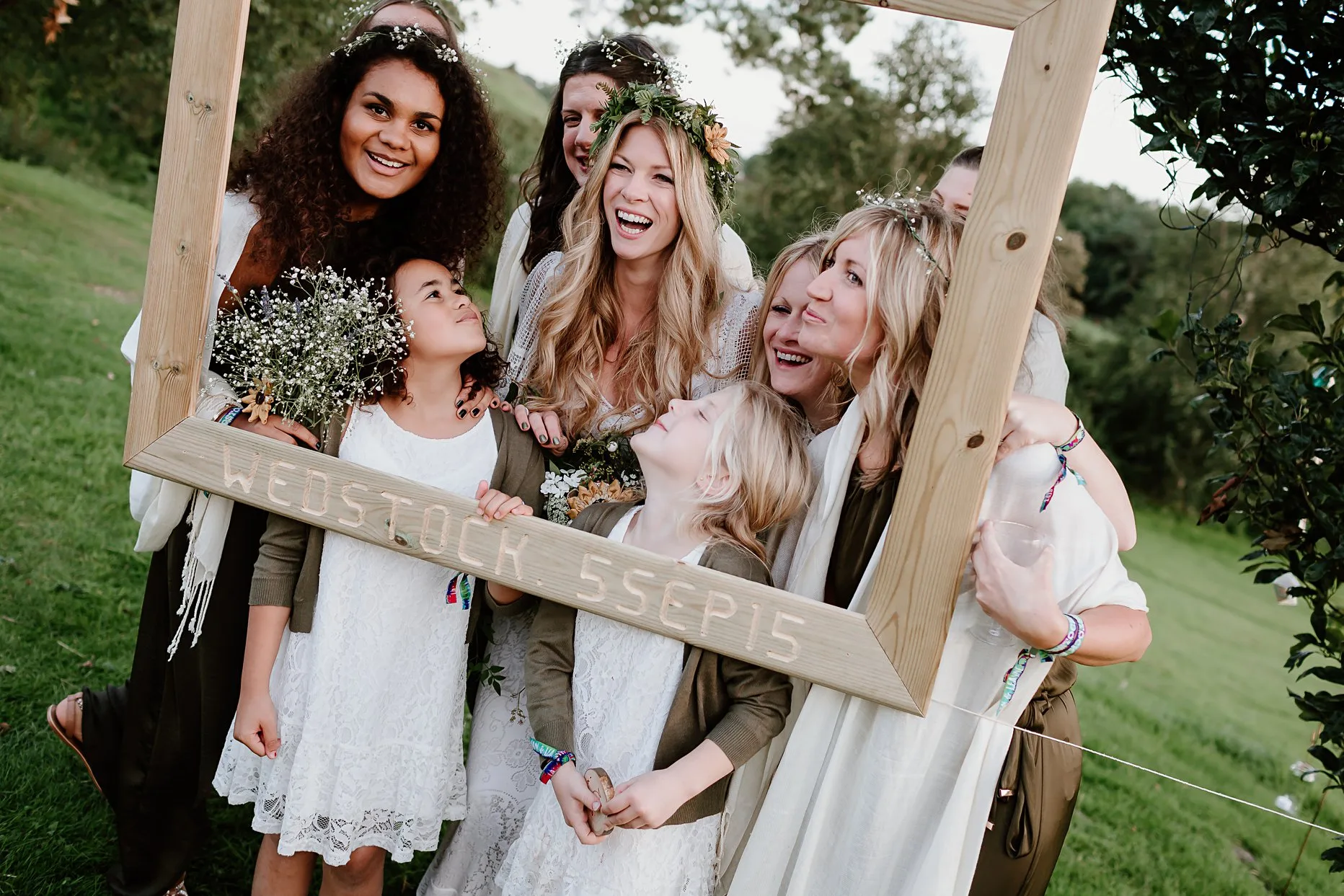 Bride and bridesmaids pose behind a large DIY wooden photo frame hung from a tree. 