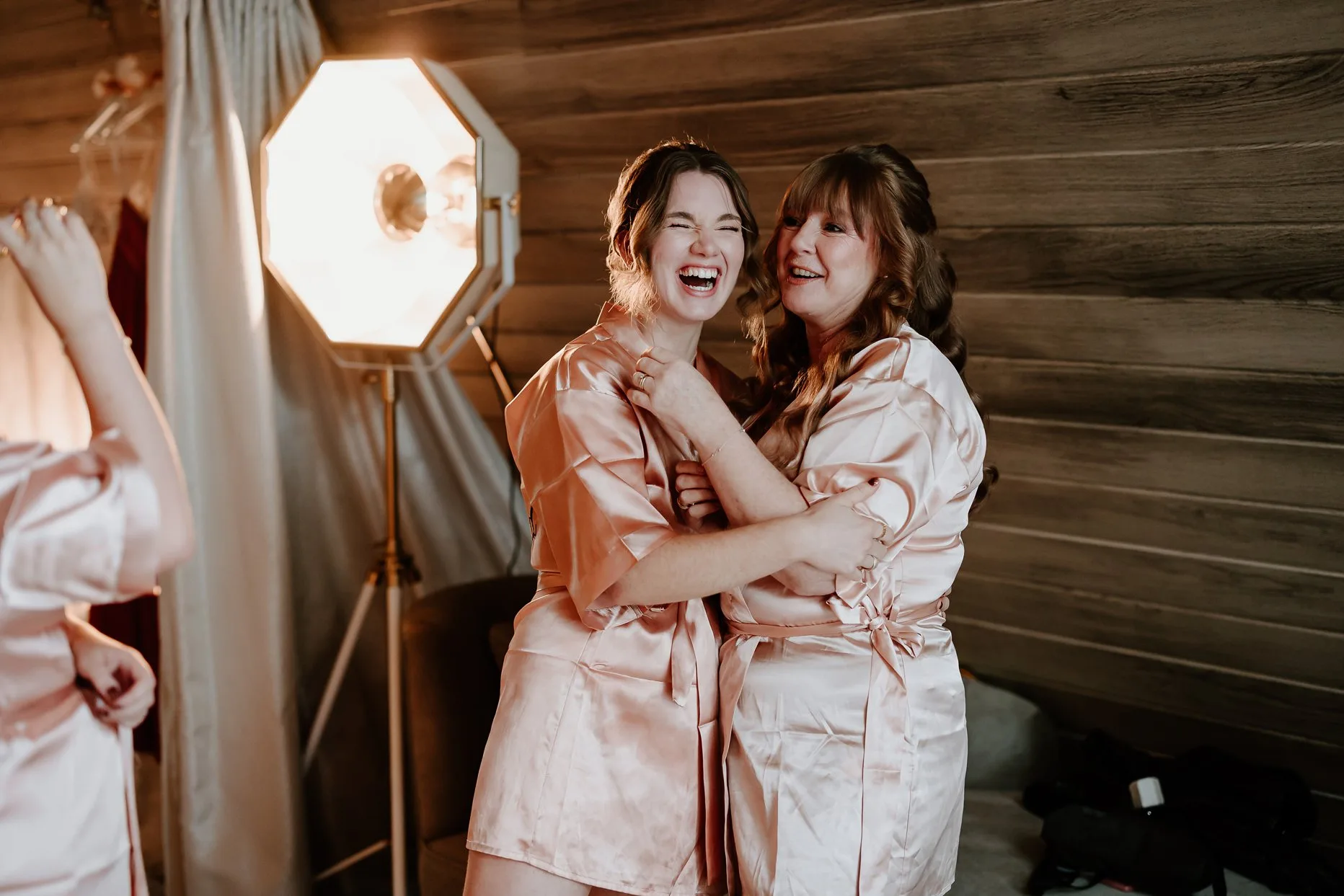 Natural photo of mother and daughter laughing on the morning of her wedding at Oaklands Grand Lodge. Both are wearing pink silk dressing gowns.