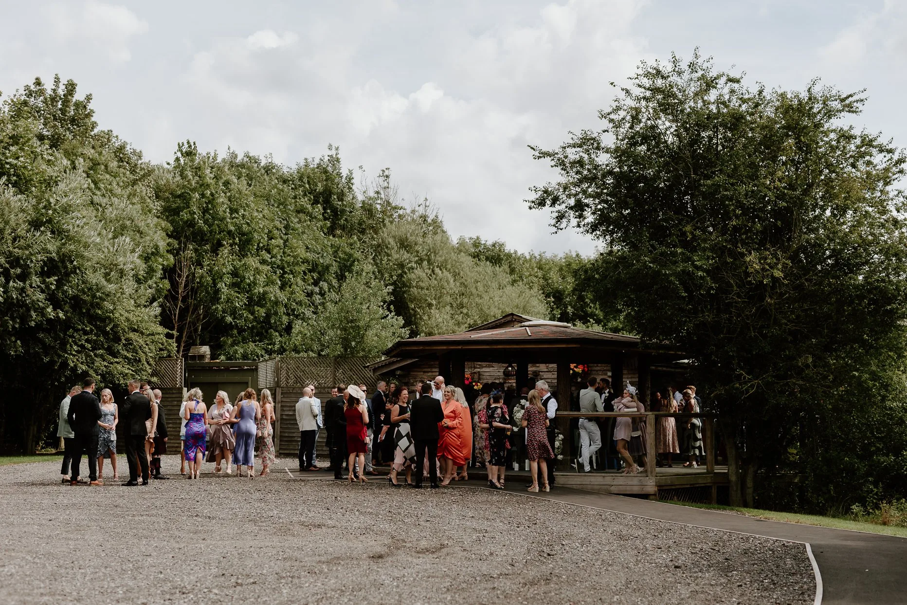 Wedding guests waiting outside the Lakeside Lodge before a wedding ceremony at Oaklands. The sun is shining and the trees surrounded the area are green.