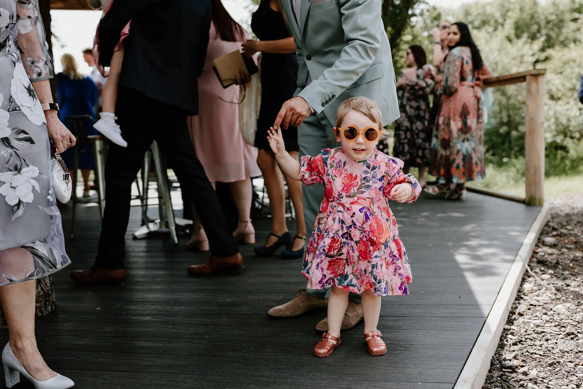 Young wedding guest wearing sunglasses stood outside waiting for the wedding ceremony to start. She is smiling at the camera and wearing a bright pink dress.