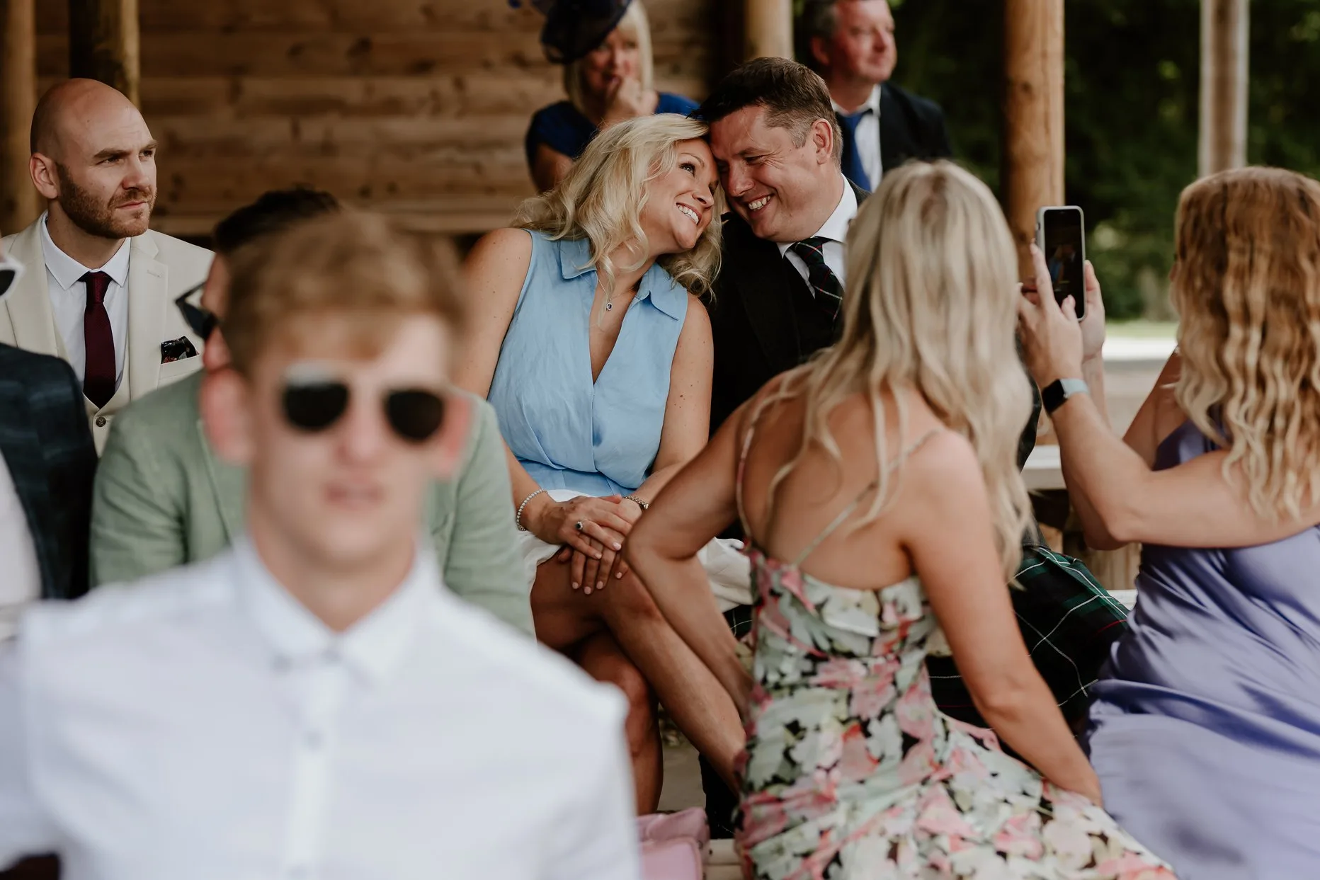 Two wedding guests smiling at each other whilst another guest take their photograph. All the guests are sat in the outdoor ceremony area at Oaklands.