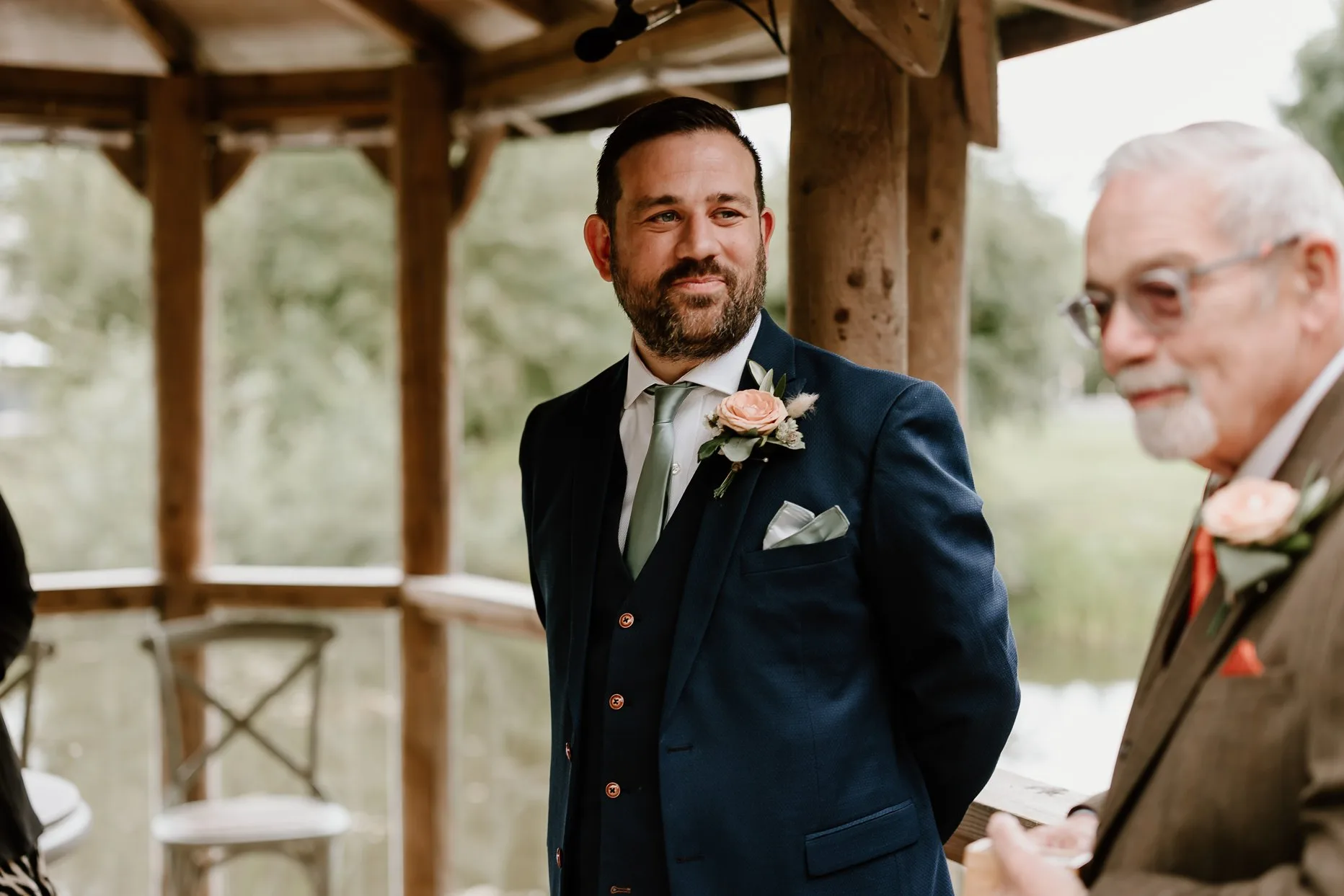 Groom waiting for his bride at the pavilion outdoor ceremony area at Oaklands. He is smiling and wearing a navy blue suit with blush pink buttonhole.