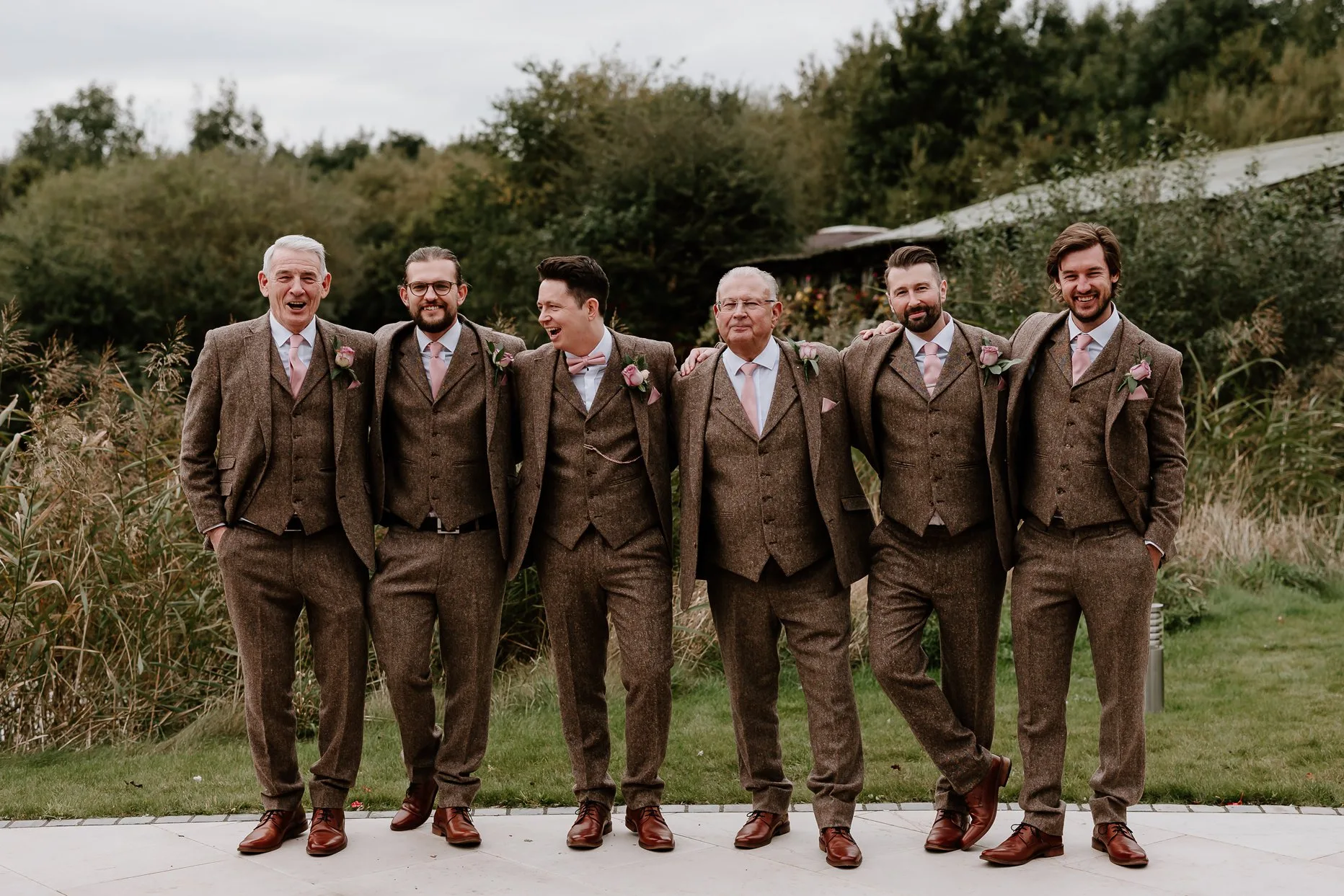 Natural photo of groom laughing and smiling with his groomsmen at Oaklands. They are all wearing matching brown tweed suits with baby pink ties and pocket squares.