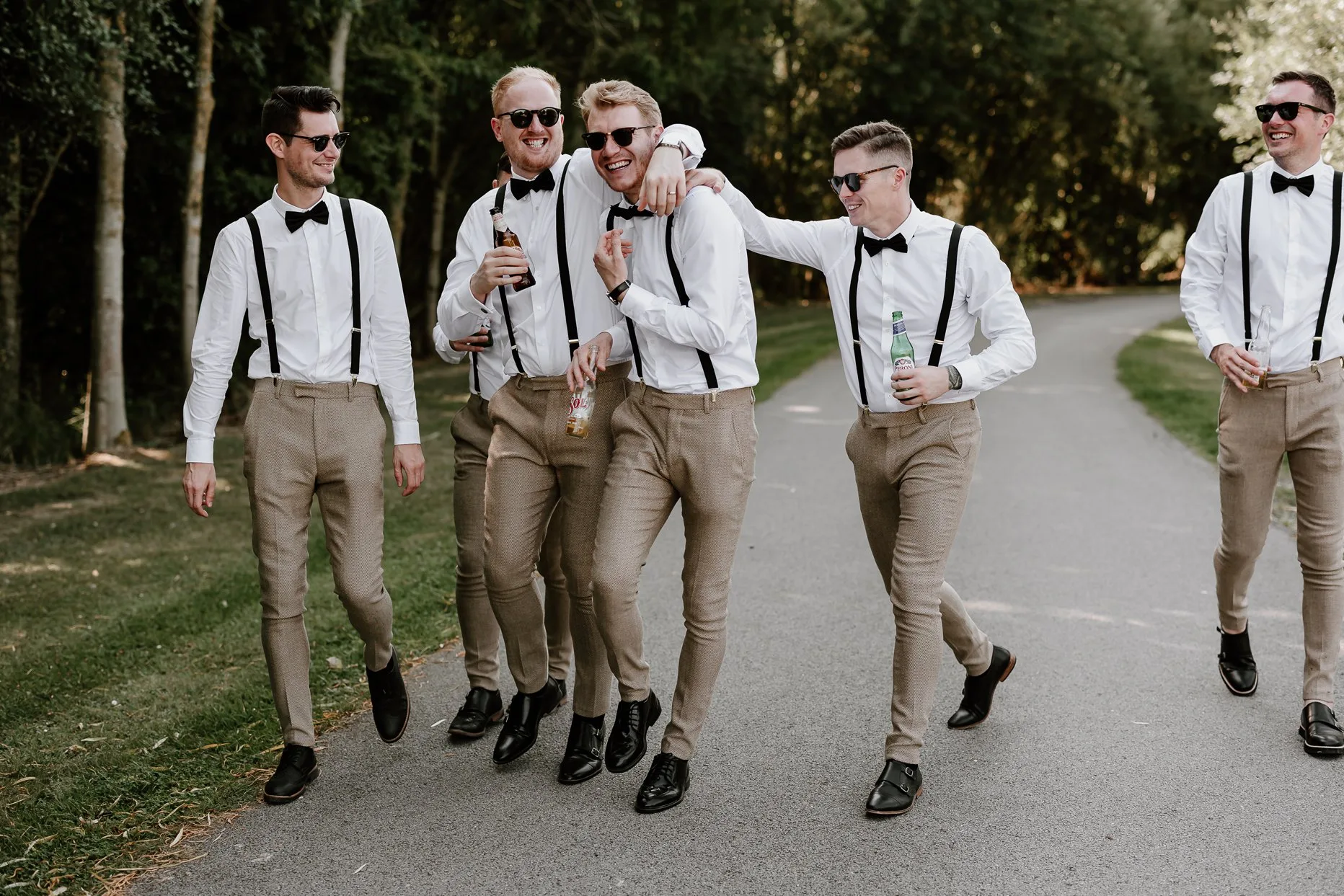 Groom and groomsmen walking towards the camera. They are wearing matching beige trousers,  black suspenders and sunglasses. They are laughing and smiling.
