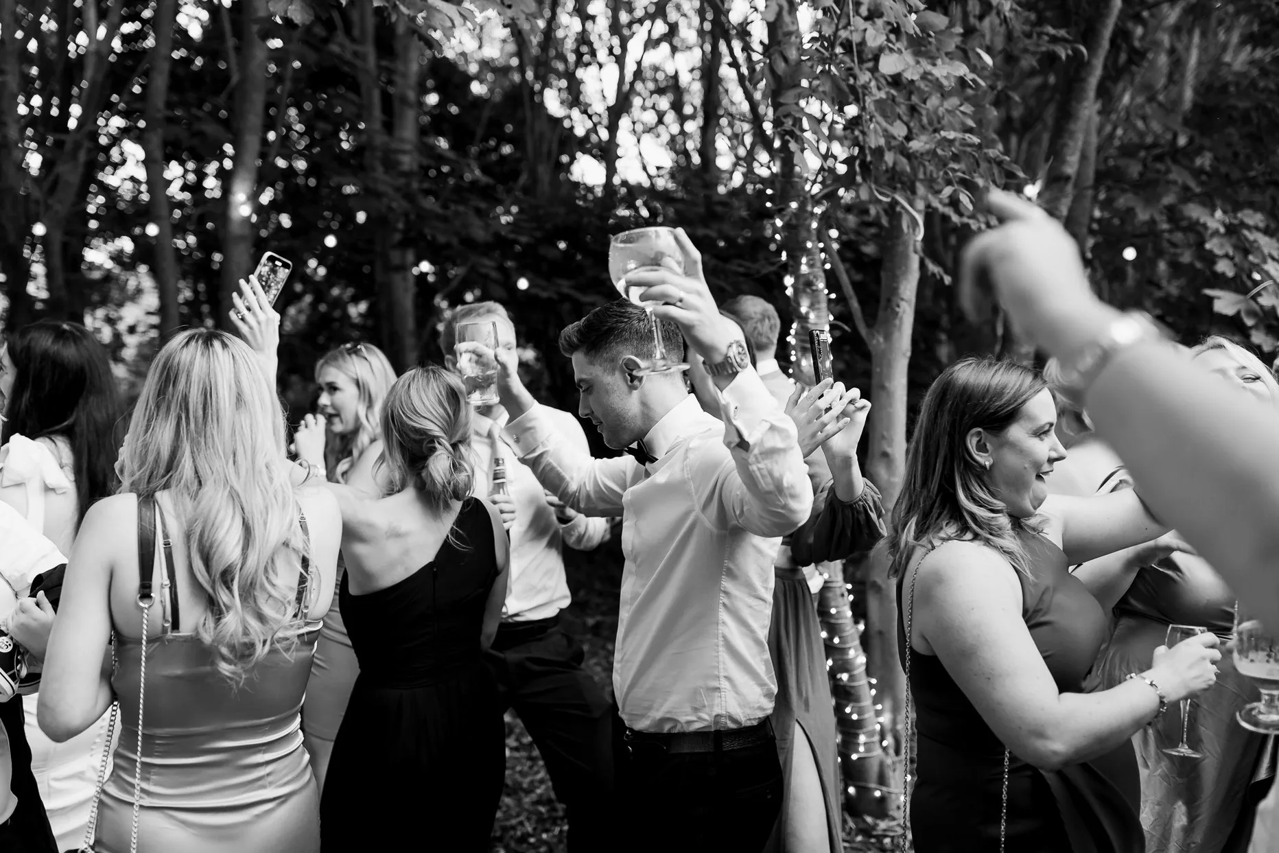 A male wedding guests dancing amongst a crowd of people. He has his hands in the air and it holding a glass of gin. 